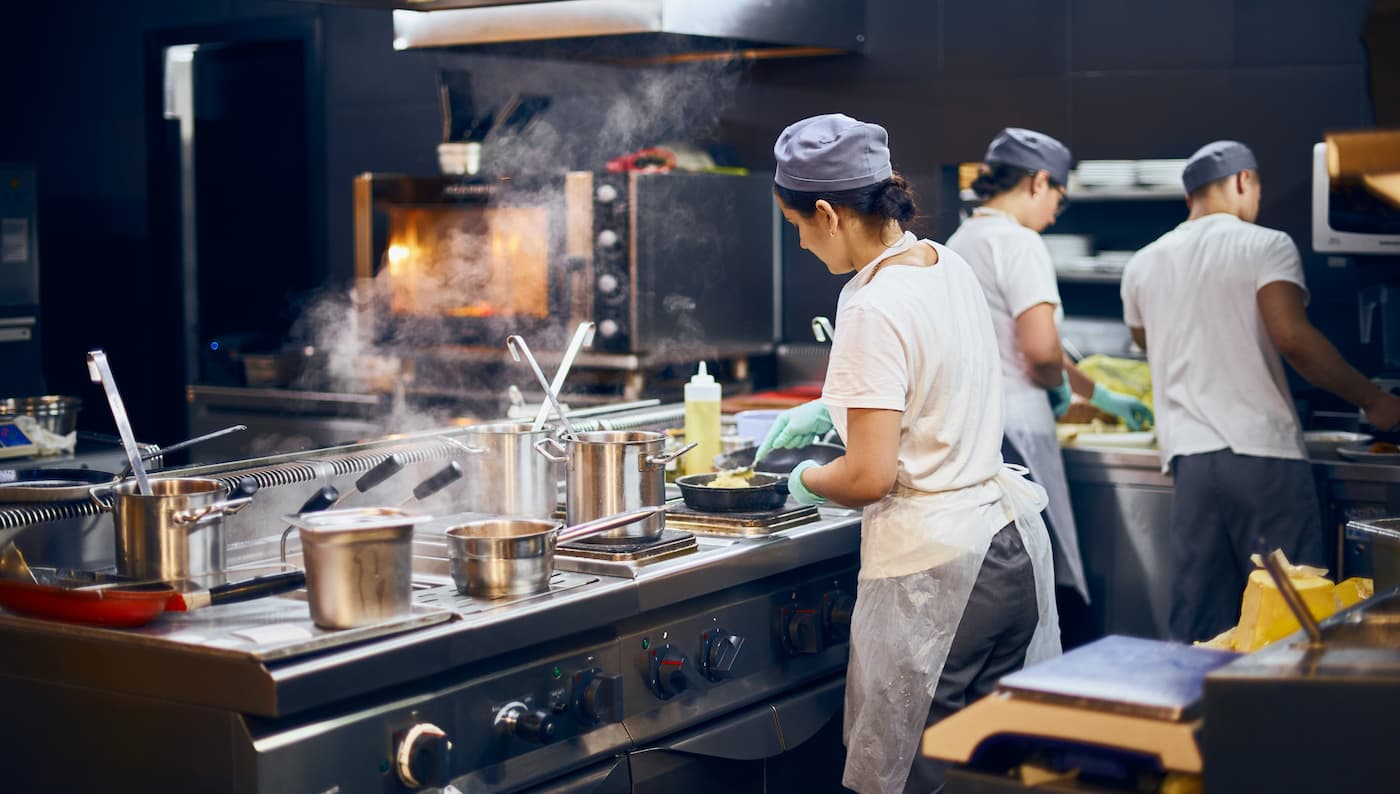 Steam rises from pots as chefs in striped aprons prep food at stove; trays, ovens, and teamwork fill busy kitchen scene.