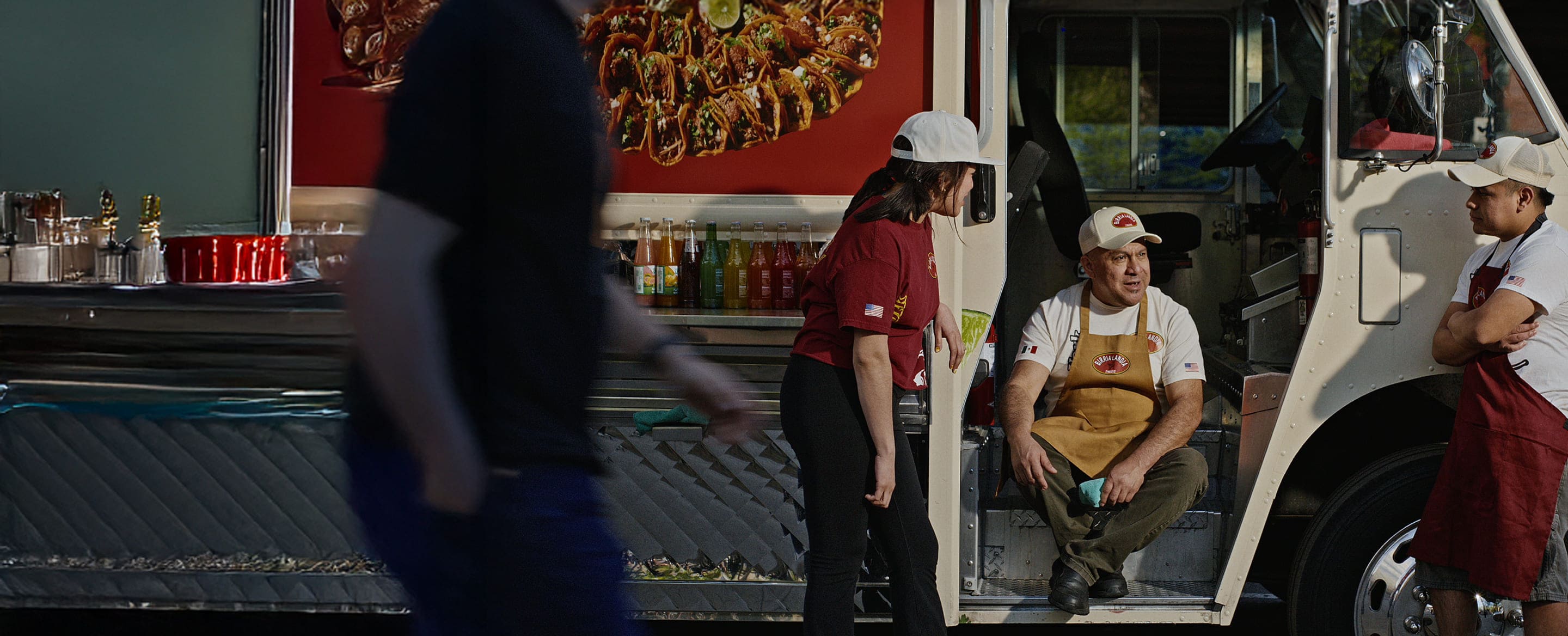 Three food truck staff near passenger door; taco image on truck, drinks and condiments on counter, blurred passerby nearby.