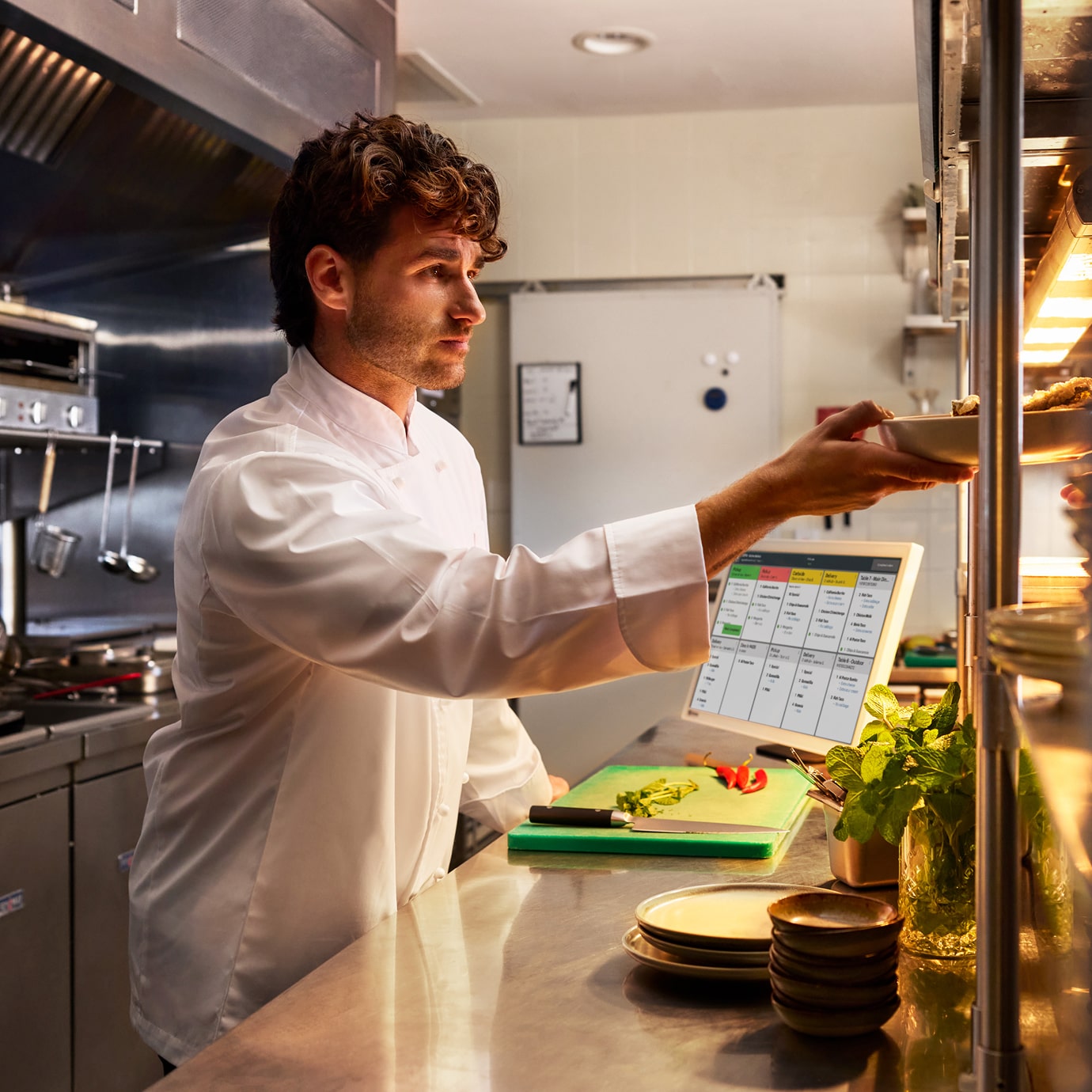 Chef places plated dish on service shelf; cutting board and kitchen display system visible in background.