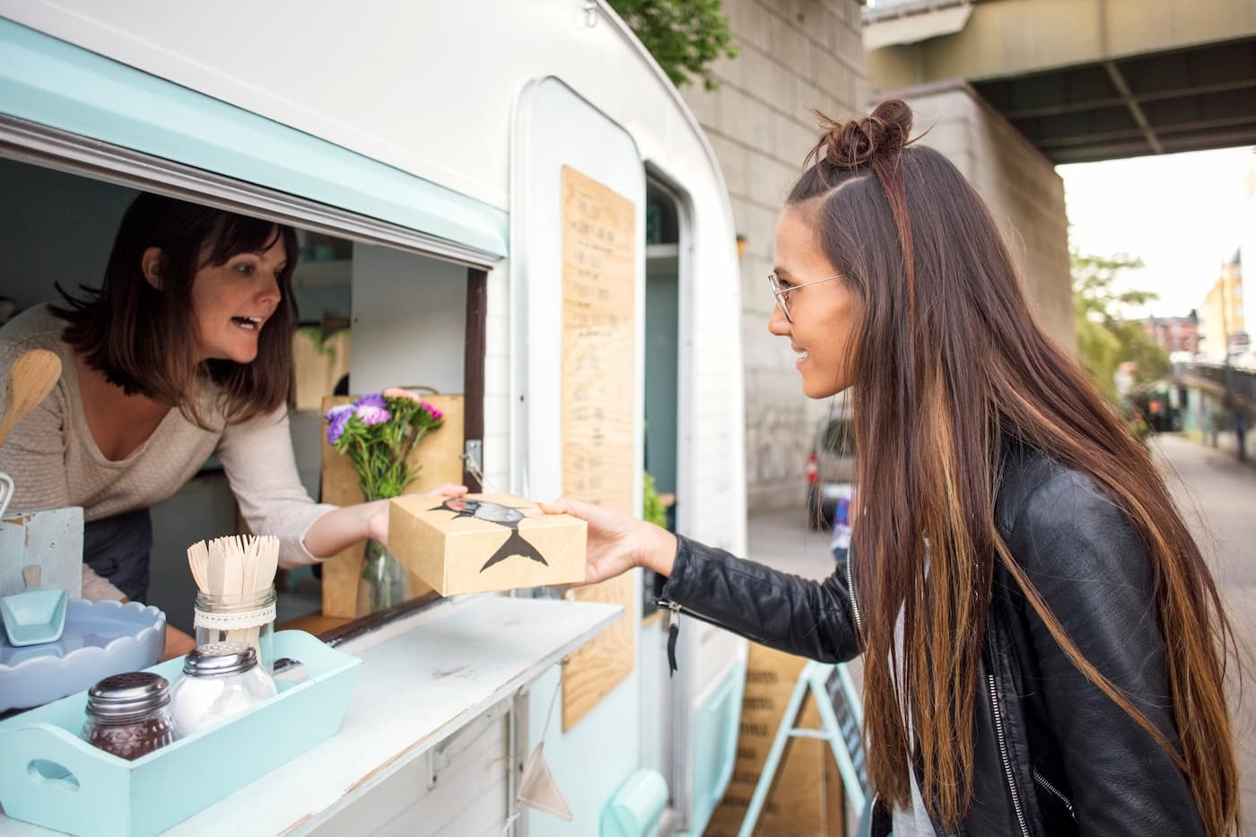 Woman in black jacket receives food from blue-and-white truck; condiments, flowers, and urban overpass visible in background.
