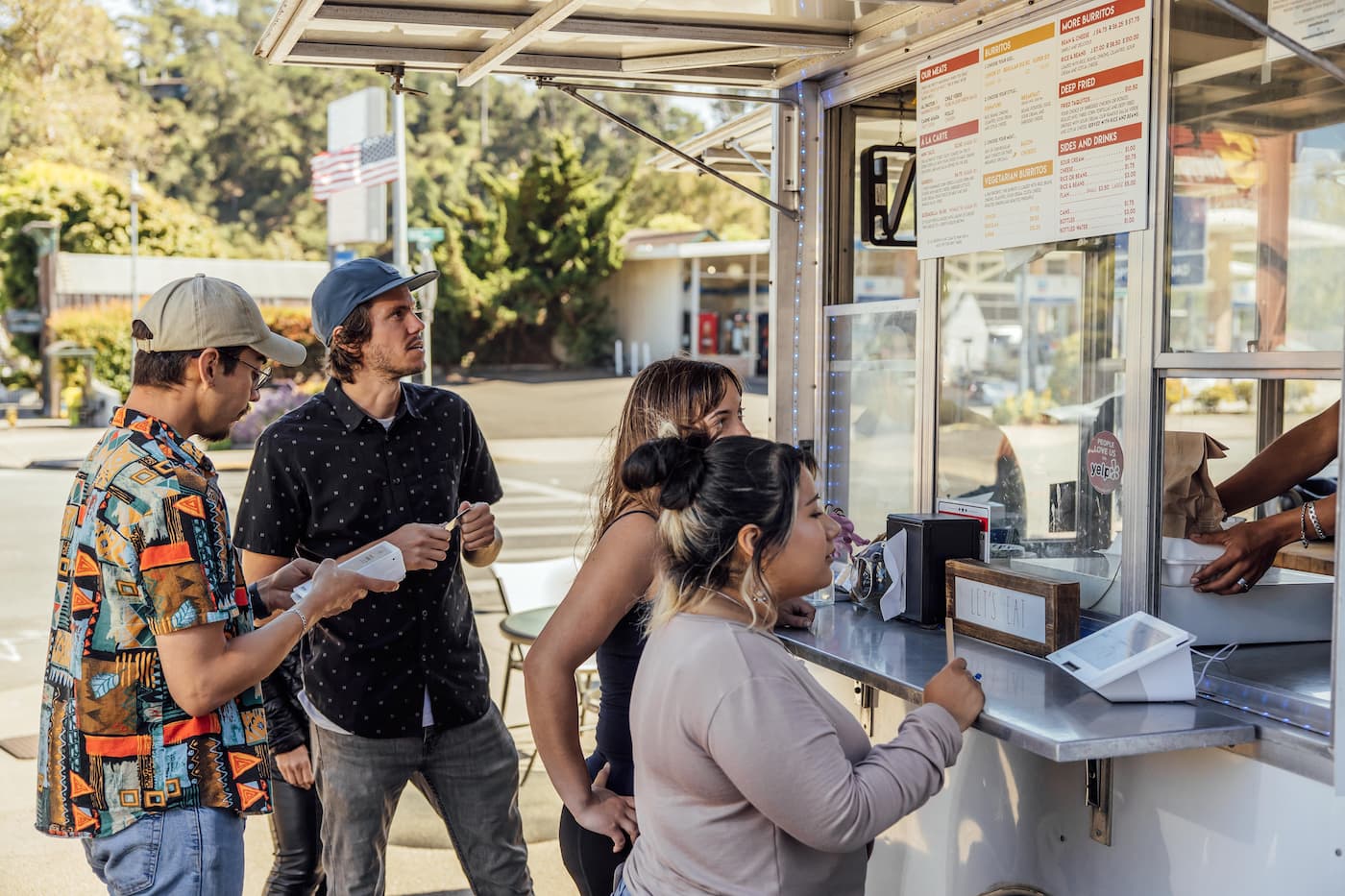 Customers at food truck: one reads menu, one orders at counter, one waits; worker takes an order using Clover Flex device.