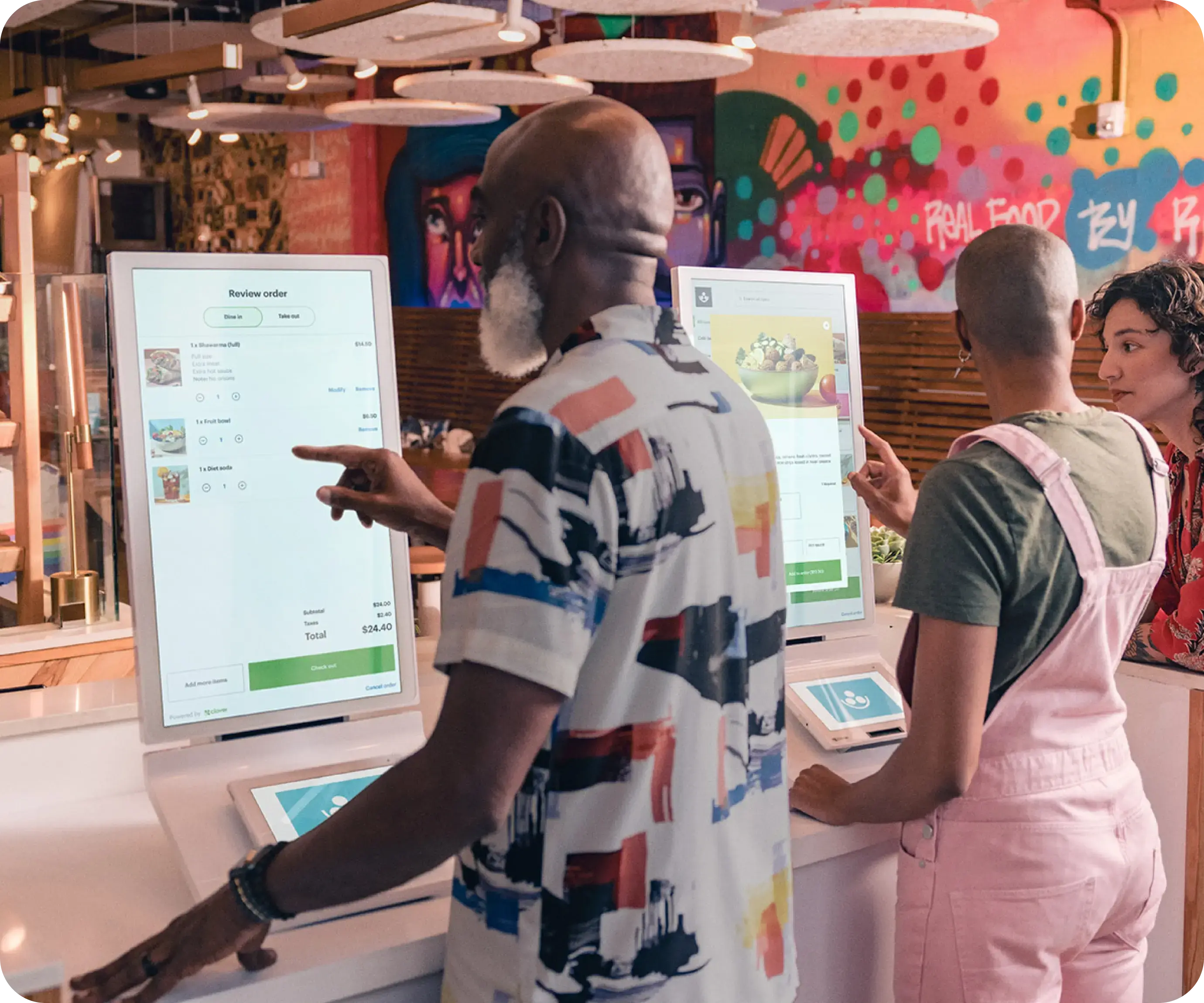 Three customers at a restaurant standing in front of Clover kiosks browsing menu items.