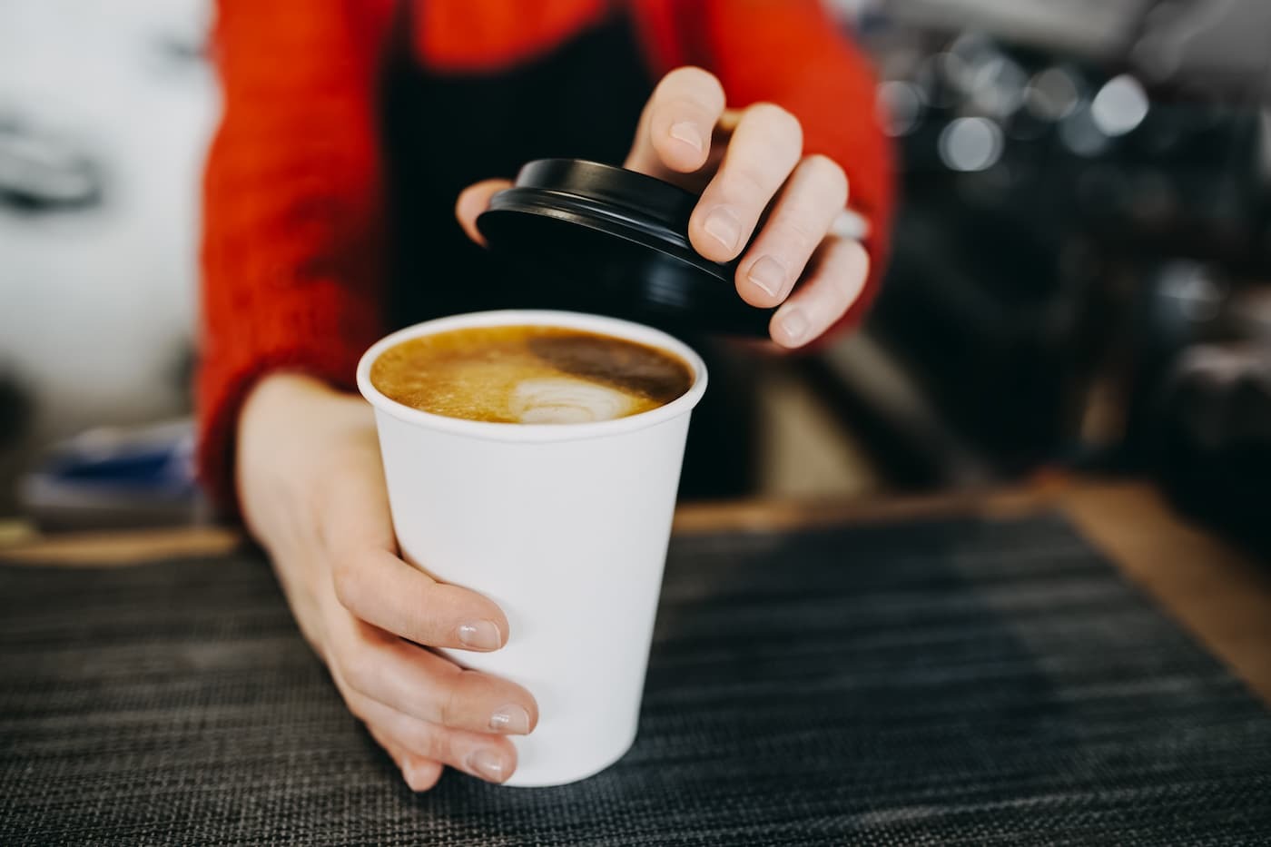 Barista wearing red sweater and apron places lid on latte with foam art; close-up of hands, blurred café background.