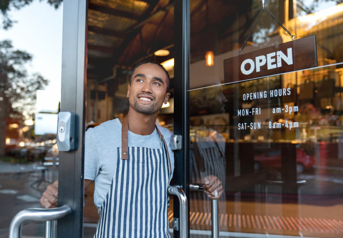Barista in striped apron holds café door open; door reads: “OPEN” with hours: Mon–Fri 8am–3pm, Sat–Sun 8am–4pm. Café visible.