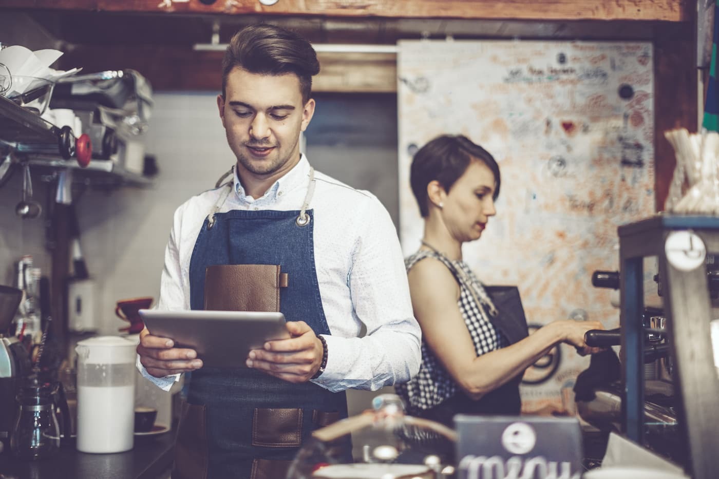 Male barista in white shirt and blue apron uses tablet; female barista makes espresso in back. Barista area in view.