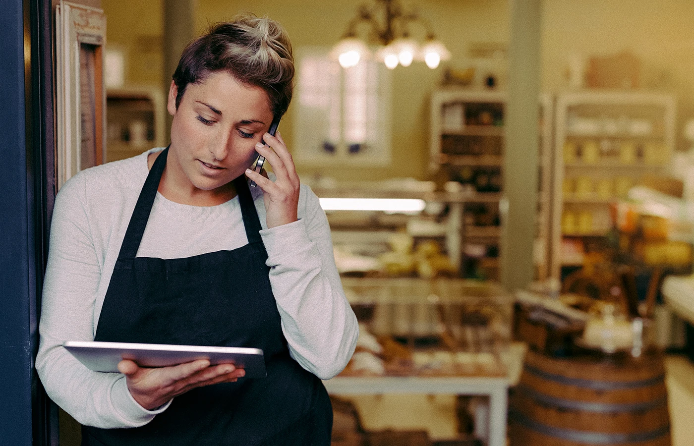 Woman standing in the kitchen of a bakery talking on a cellphone while using a tablet 