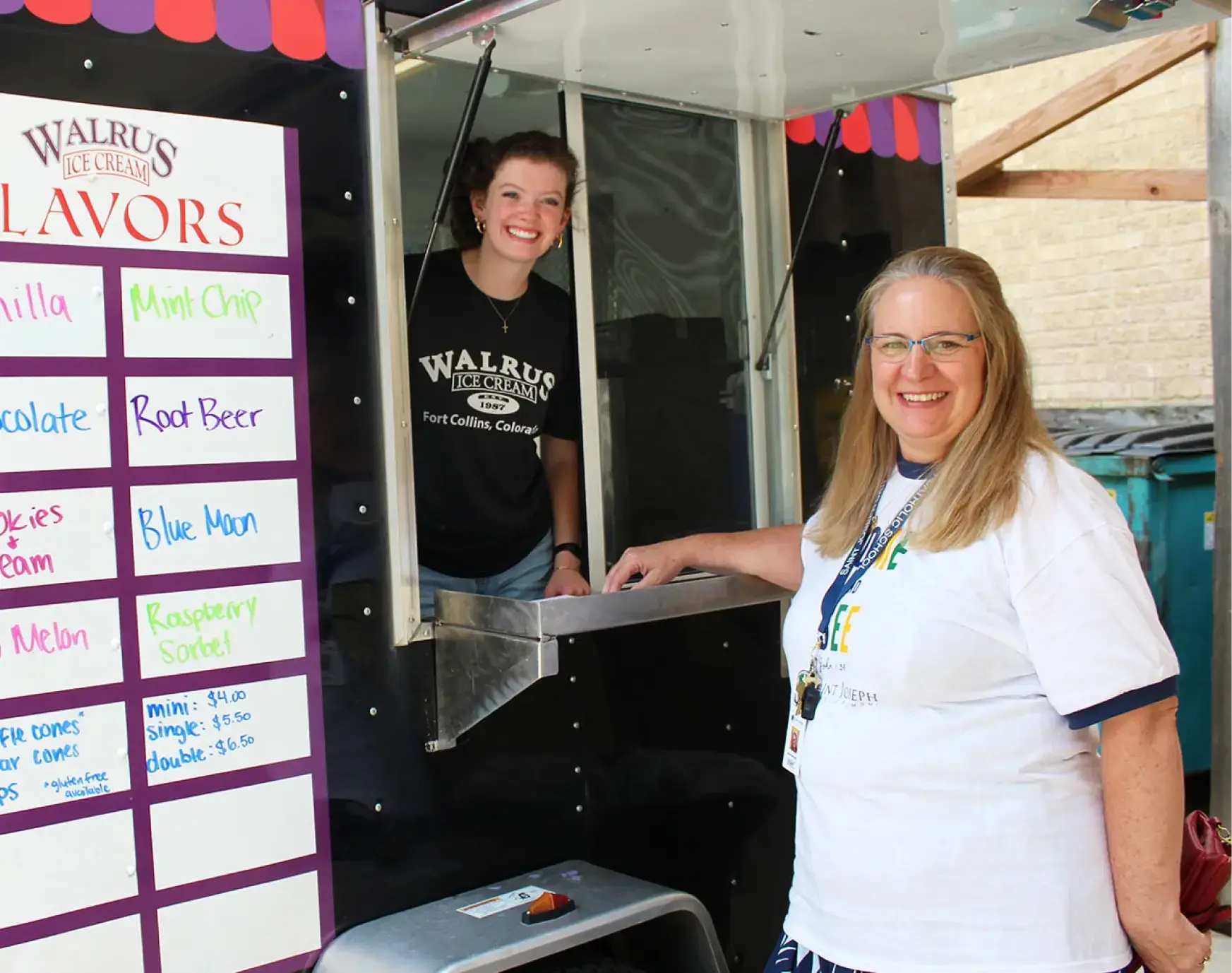 Woman standing inside an ice cream truck while a customer stands outside smiling and waiting to be served. Ice Cream flavors are presented on a board outside of truck.