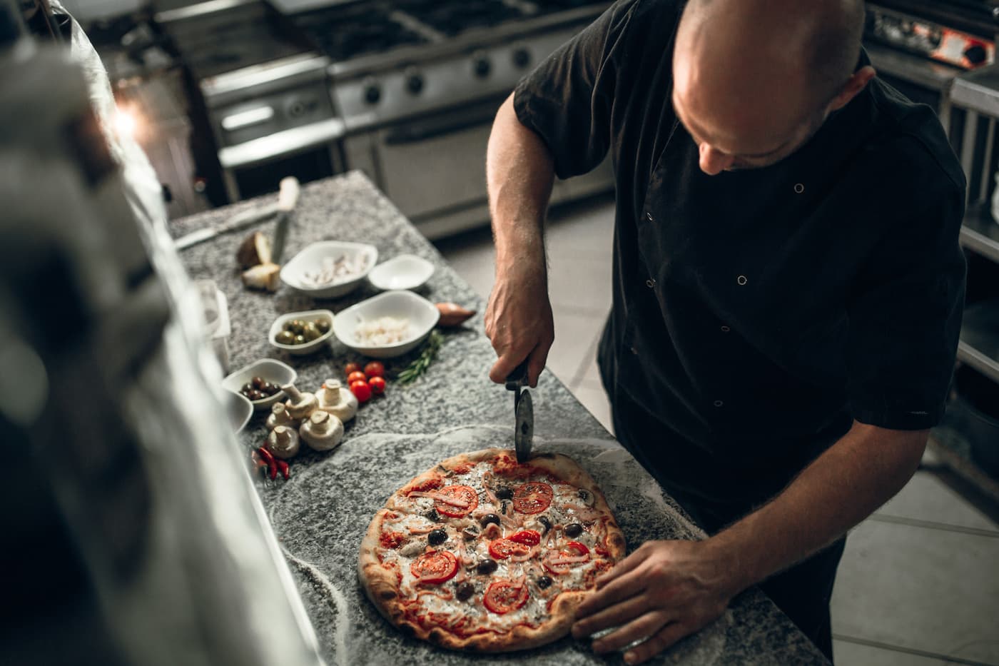Chef slices pizza topped with tomato, mushrooms, sausage in kitchen; fresh ingredients in bowls nearby on stone counter.