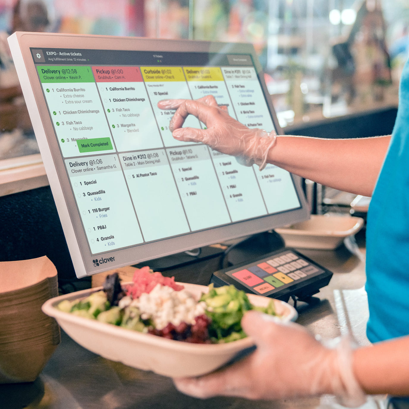 Food service worker at a Mediterranean restaurant holds a completed to-go bowl while marking it as done on the KDS.