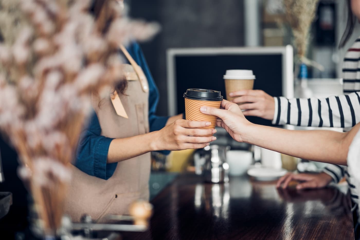 Barista in beige apron hands coffee to customer at café counter; another customer in striped shirt reaches holding coffee.
