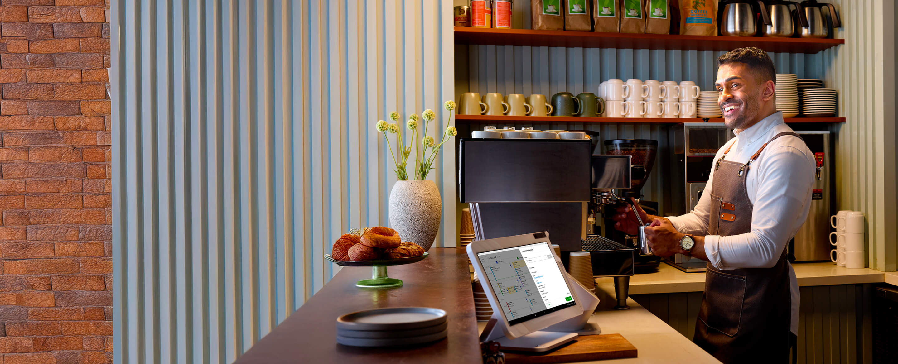 Barista in white shirt and brown apron smiles while using espresso machine; doughnuts, clover station solo, and mugs visible.