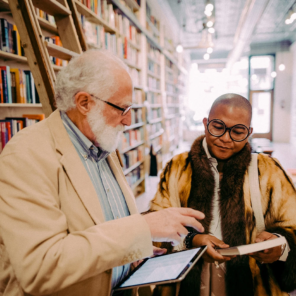 Man holding tablet points to book that woman is holding.