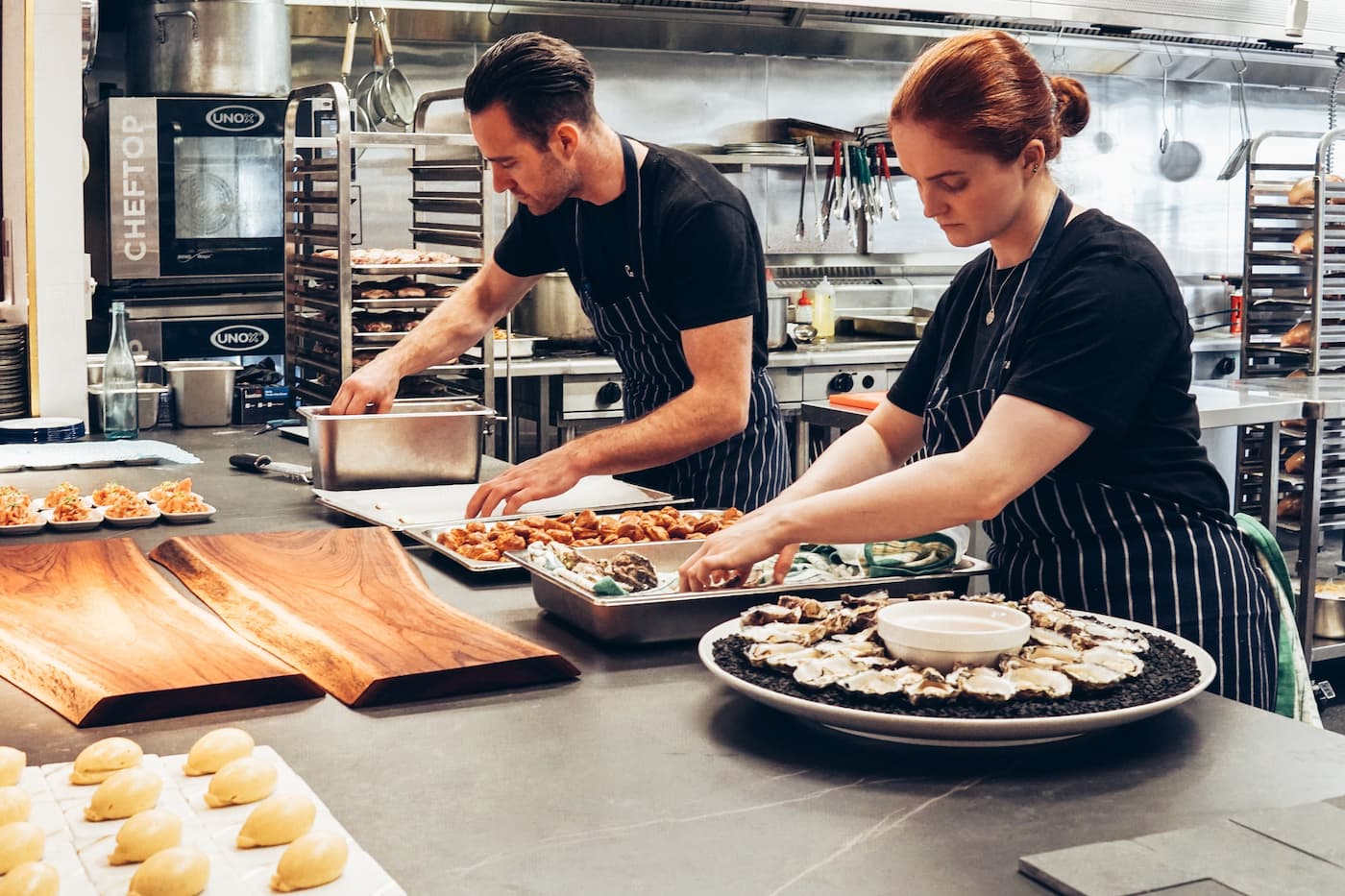 Two chefs in striped aprons prep food at stainless counter; oysters and sauce on serving boards; busy kitchen in background.