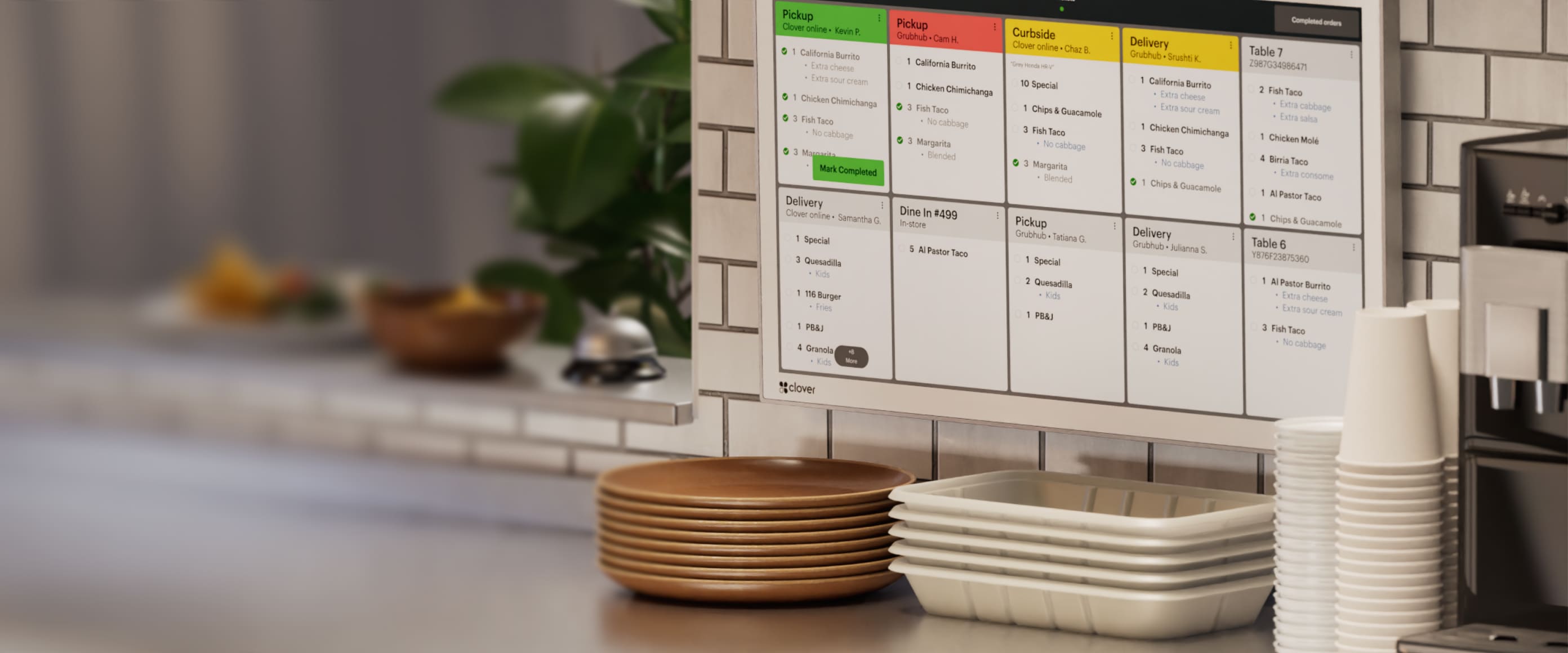 KDS screen with orders by table/status; plates, cups, water dispenser; blurred service bell, chips, food, and plant visible.