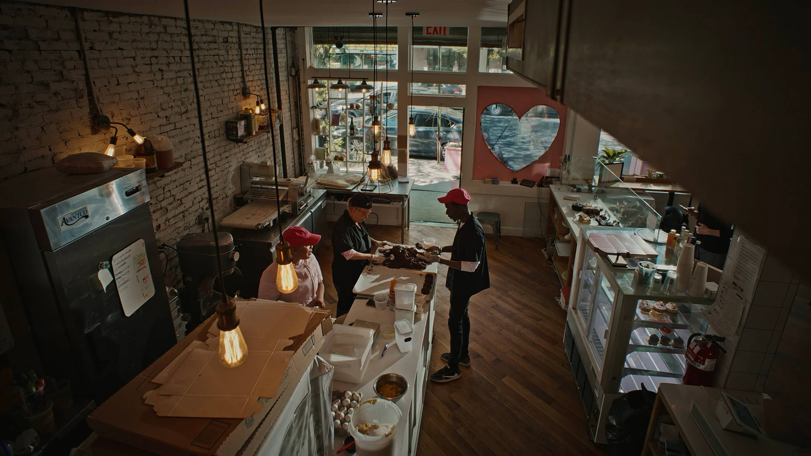 Workers are preparing pastries in a warm, rustic bakery setting; brick wall, heart window, baking equipment visible.