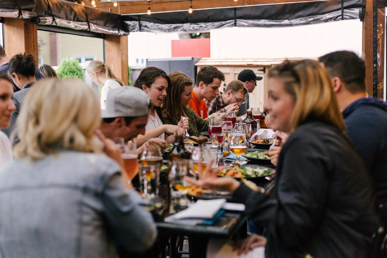 Large group of diners sitting at long, wooden tables on an outdoor patio; string lights hung, diners enjoy food and drinks.