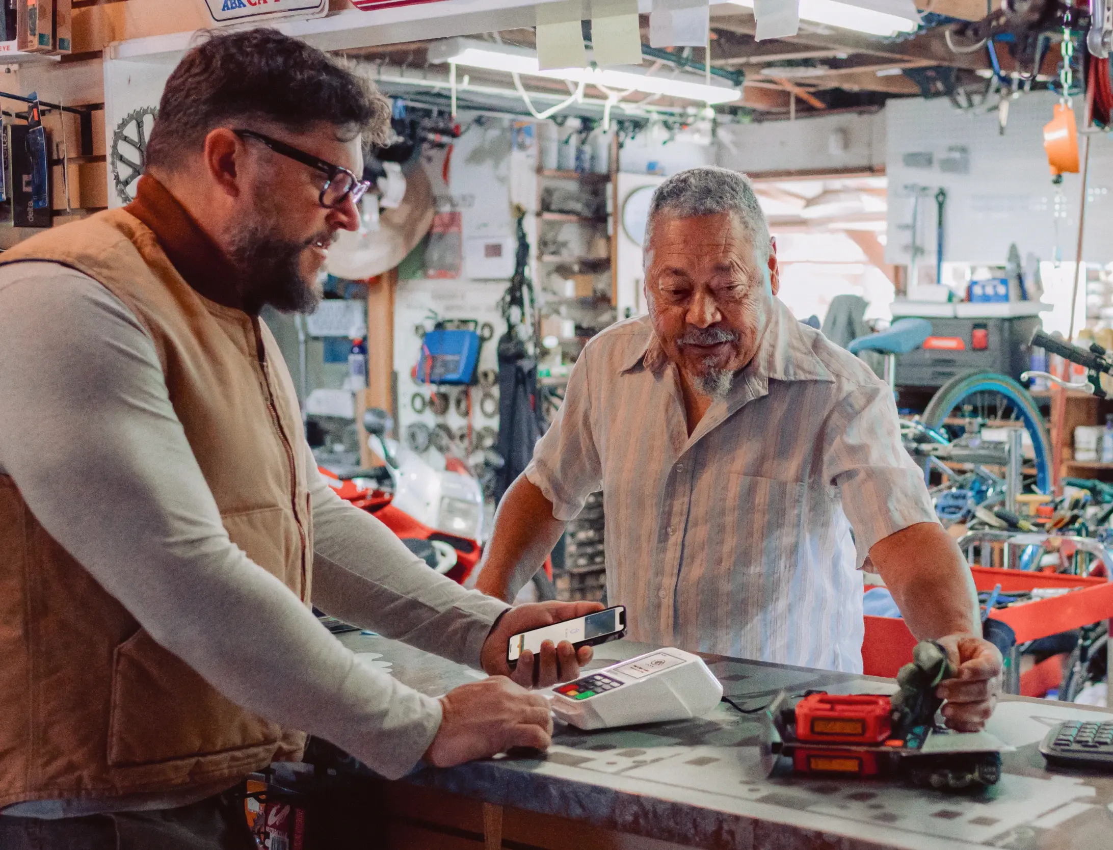 Older man in a bicycle repair shop using tap-to-pay on a Compact while shop owner touches an item the customer payed for.