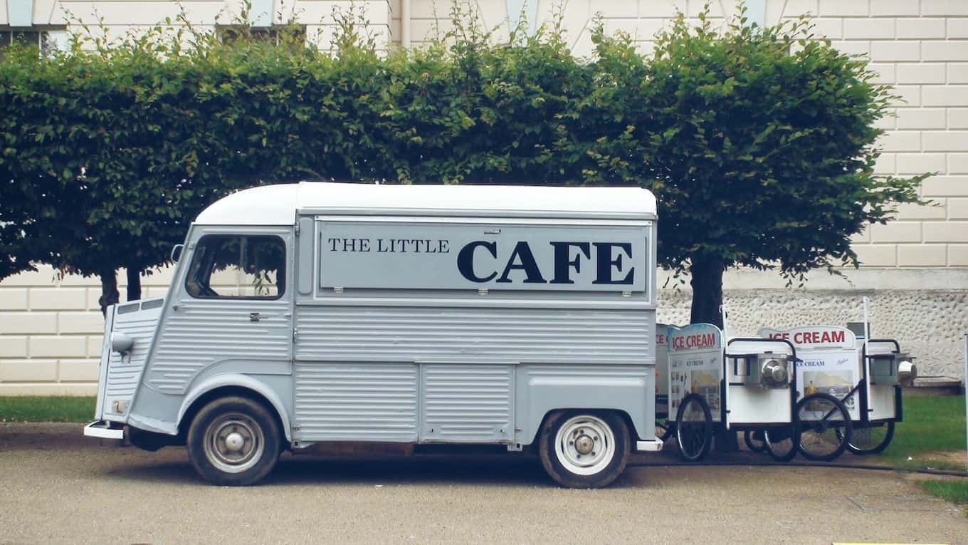 Light gray, old, small food truck labeled “The Little Cafe” parked in front of trees; ice cream wagons parked behind truck.