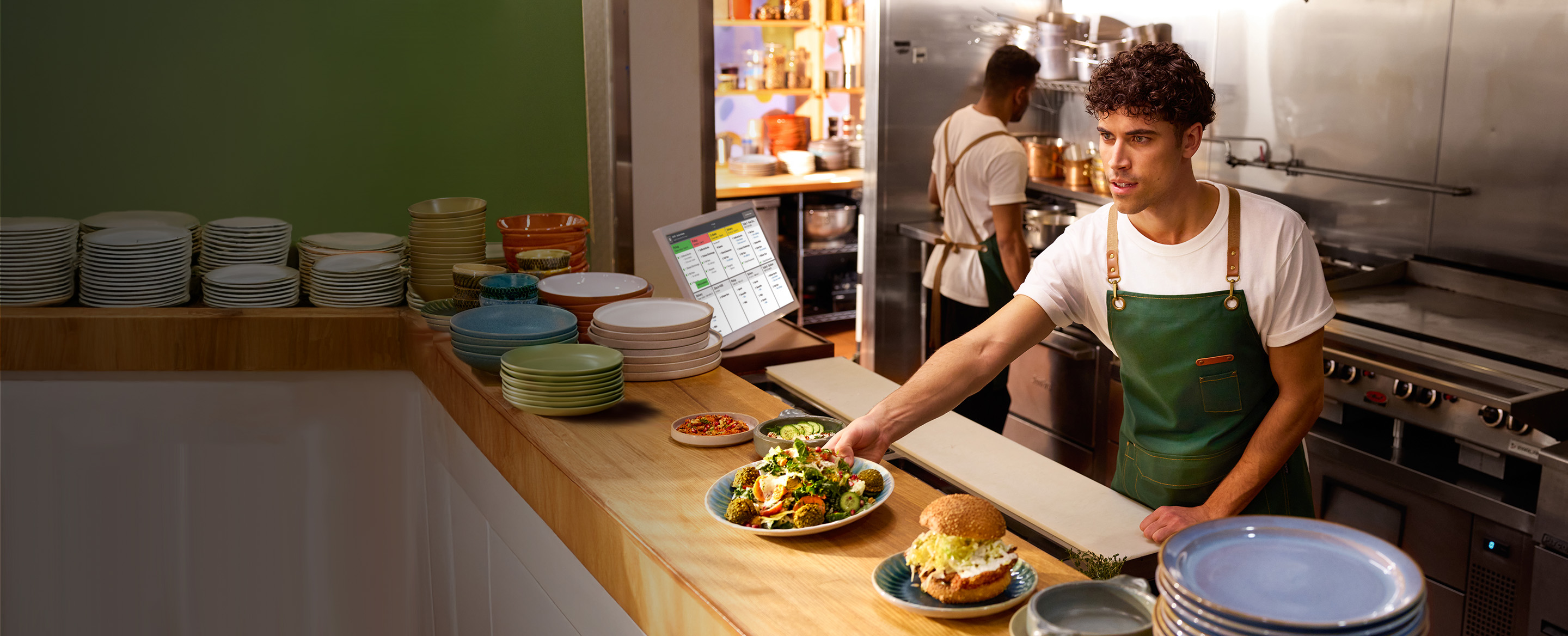 Chef places plated dish on wooden counter for service; KDS screen in background with order instructions in kitchen setting.