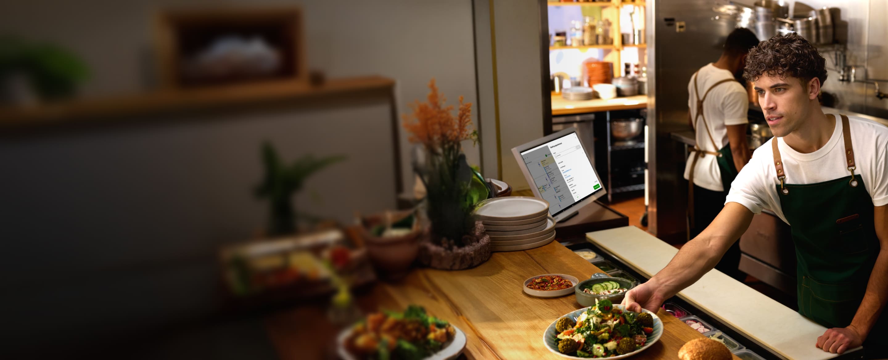Chef places plated dish on wooden counter for service; KDS screen in background with order instructions in kitchen setting.