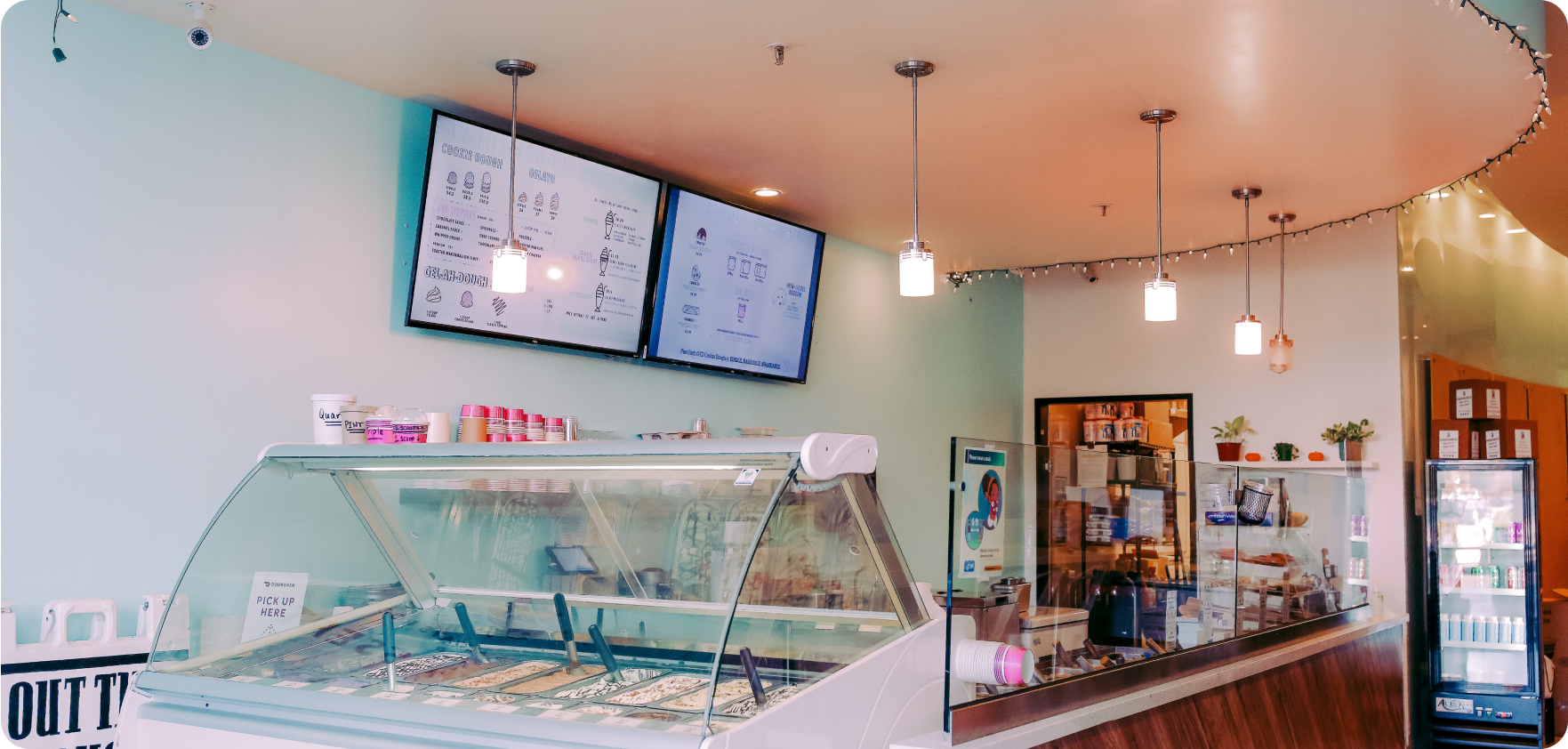 Dessert place featuring gelato in a glass case, a menu featured on two televisions, an assortment of cookie dough, and cookies in a glass case.