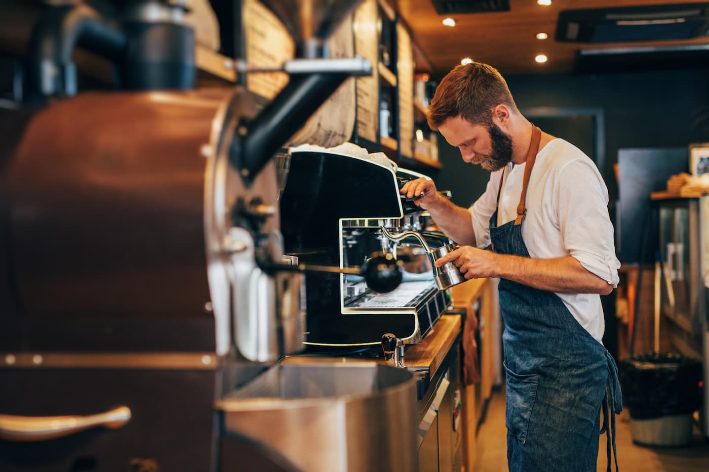 Barista in white shirt and blue apron steams milk at espresso machine; coffee roaster and tools visible in warm café setting.