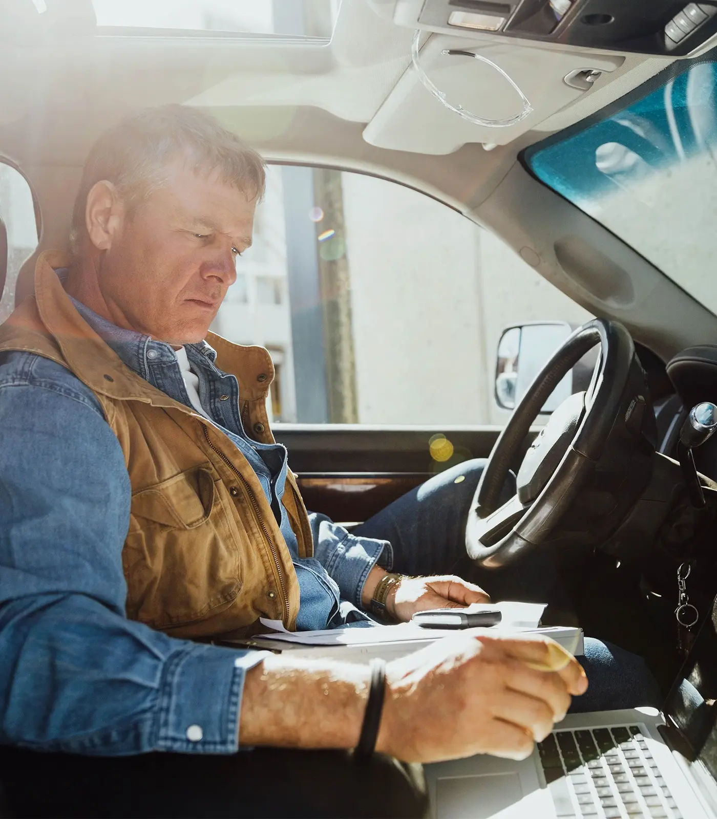 Contractor sitting in a truck holding a binder and papers in his left hand, while he uses his right hand to work on a laptop.