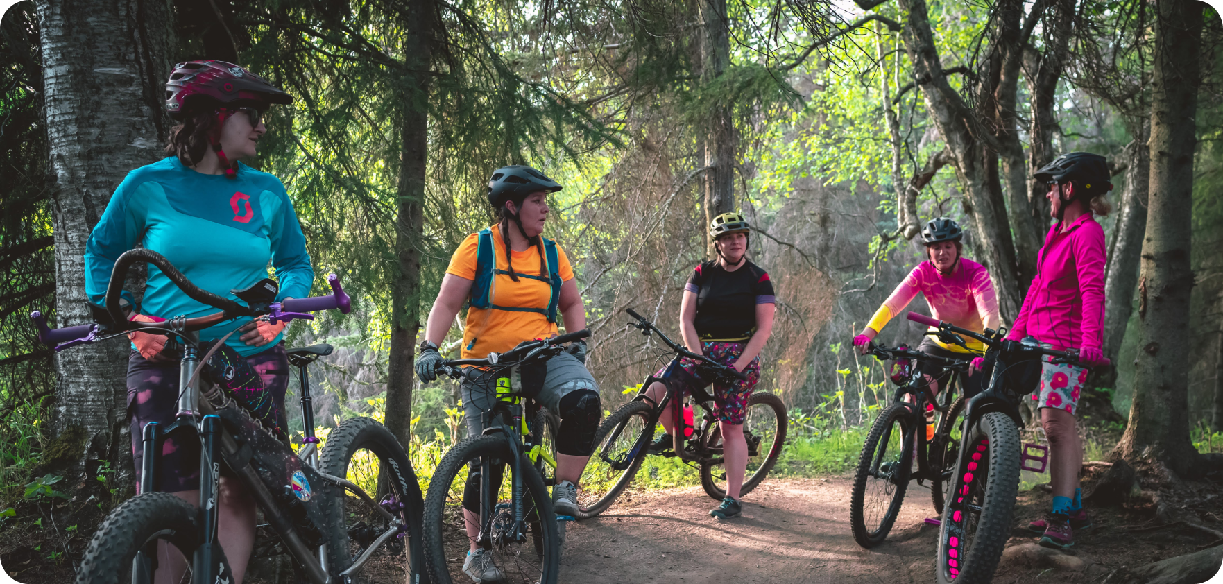 Group of five women on a bike trail in the wilderness wearing colorful gear while standing next to their mountain bikes. 