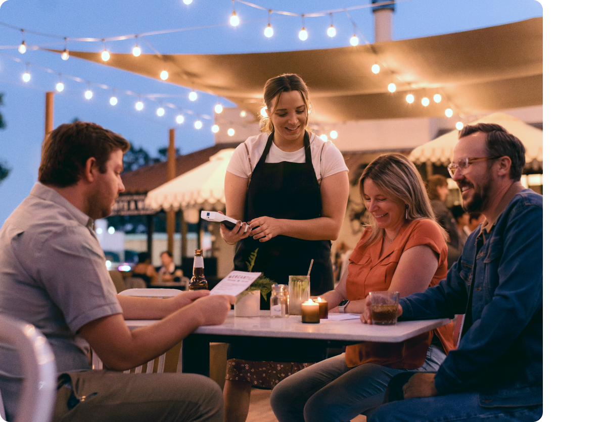 Female server smiles holding a Flex Pocket while taking diner orders at an outdoor restaurant under string lights at dusk.
