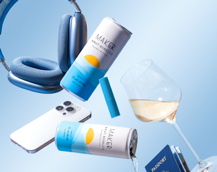 Two Maker Wine Brut Bubbles cans of wine floating in air alongside a glass of wine, a passport, a phone, and a pair of headphones.