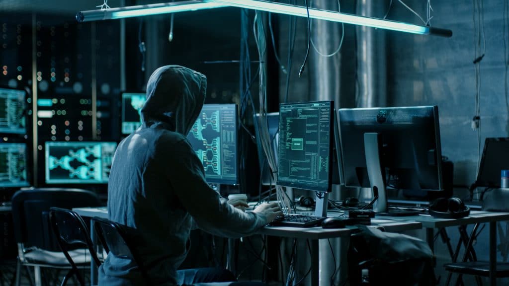 a hooded person typing on a computer in a shifty looking setting