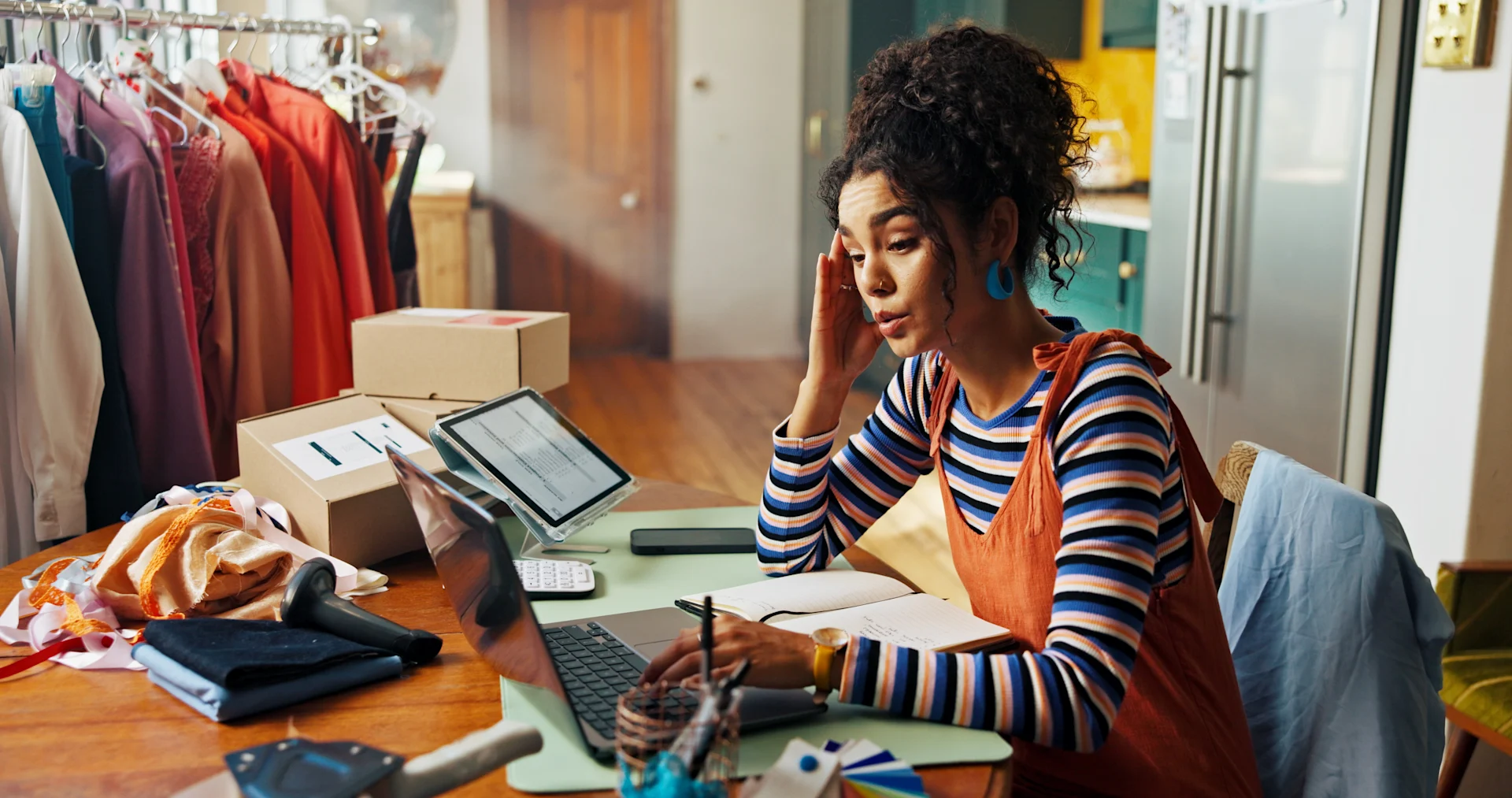 Female presenting person surrounded by clothes and boxes, working on a laptop.