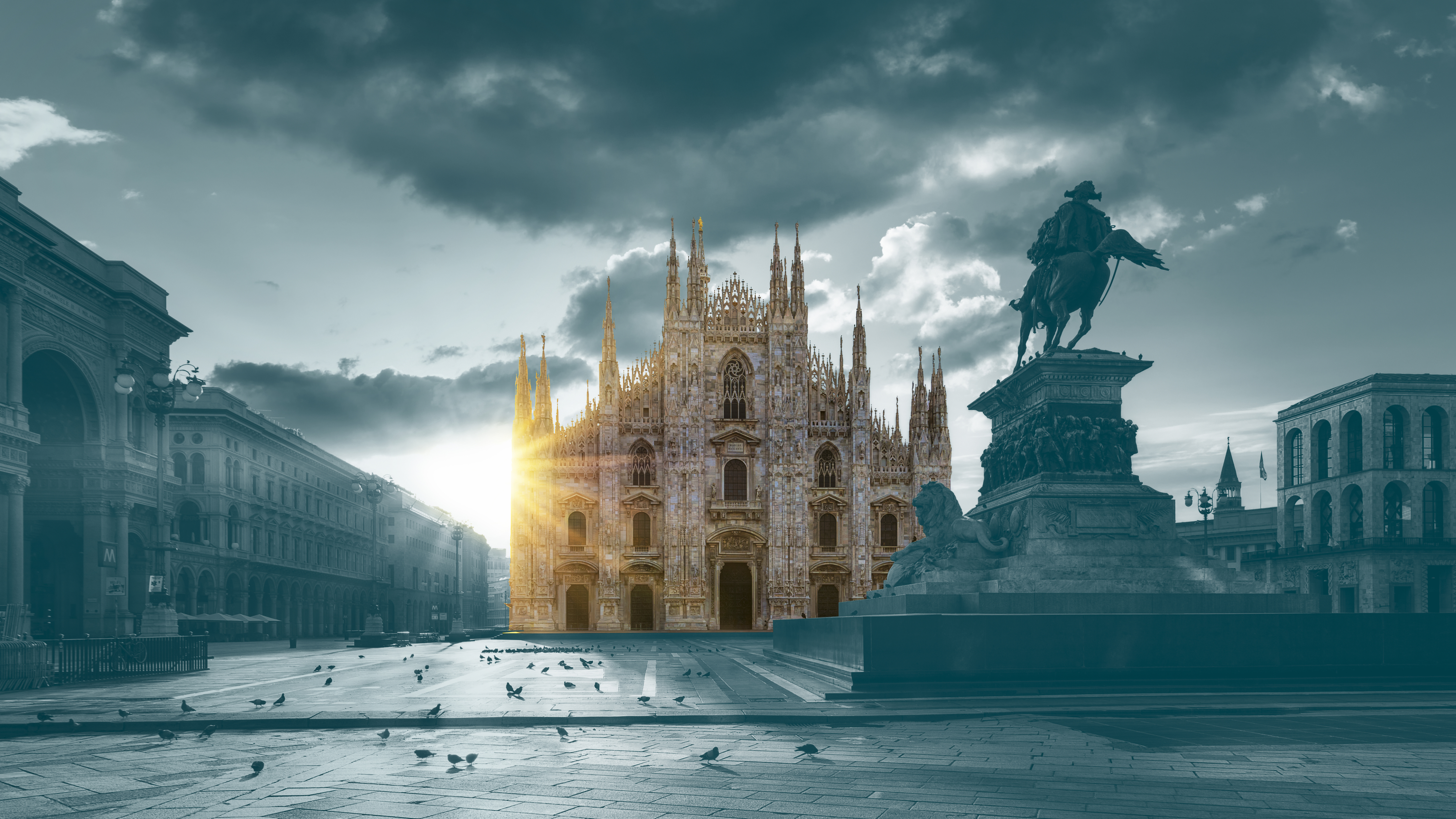 A dramatic view of Milan’s Piazza del Duomo at sunrise, with the sun illuminating the façade of the Milan Cathedral while the surrounding square and statue appear in cool, muted tones.