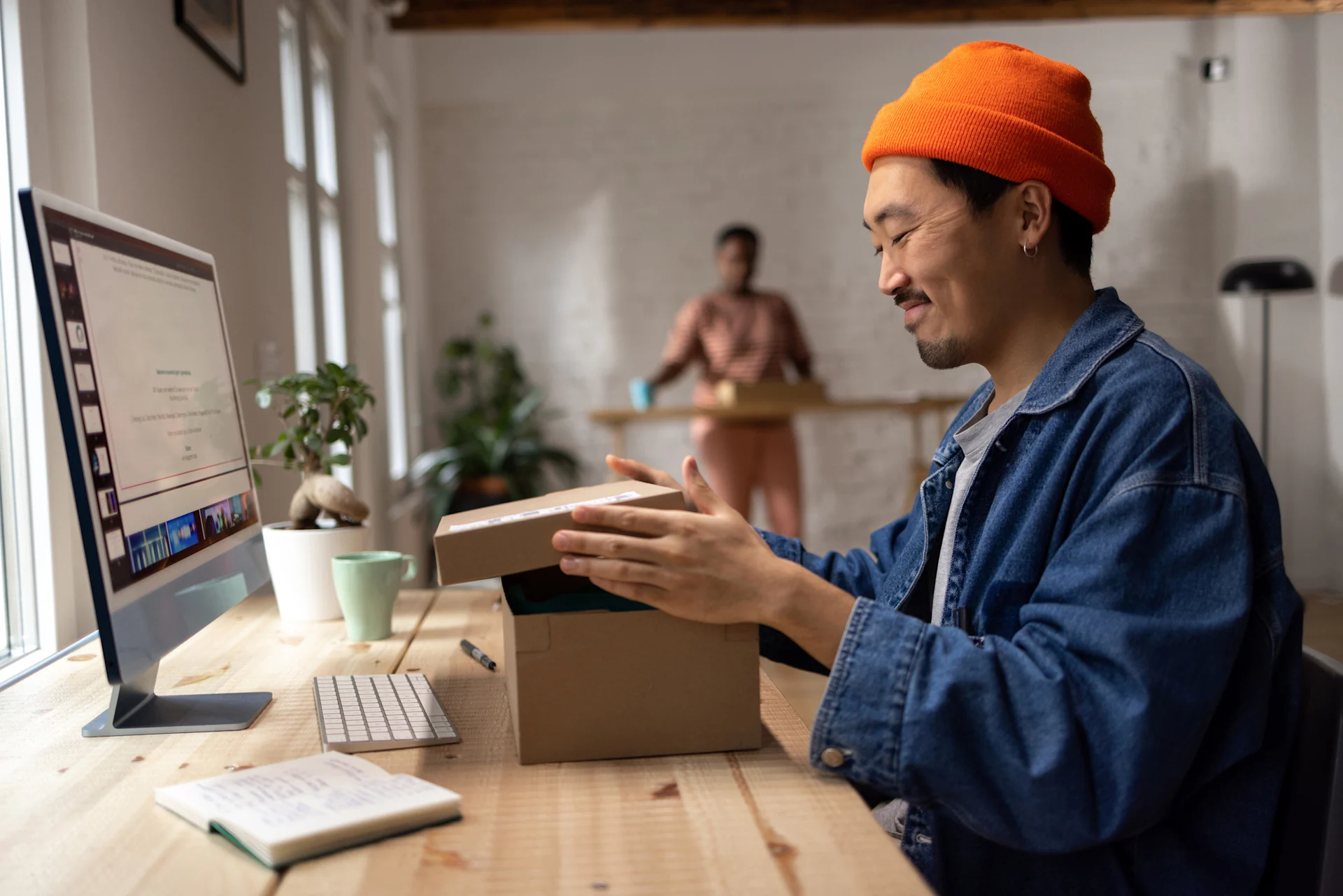 Male presenting in orange beanie smiles whilst assembling parcel box in front of their computer.