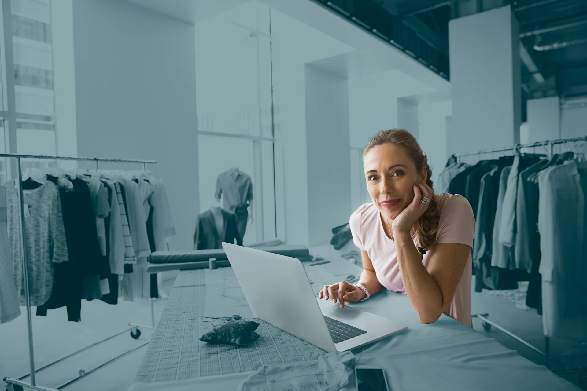 A woman sitting at a table in a clothing studio or showroom, leaning on her hand while working on a laptop, with racks of garments visible in the background.
