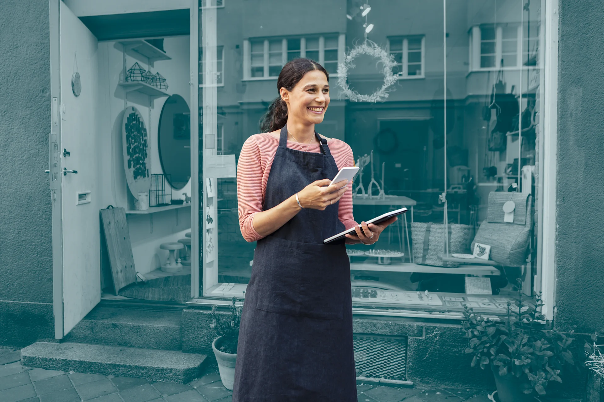 Woman in apron standing outside business with pad and phone.