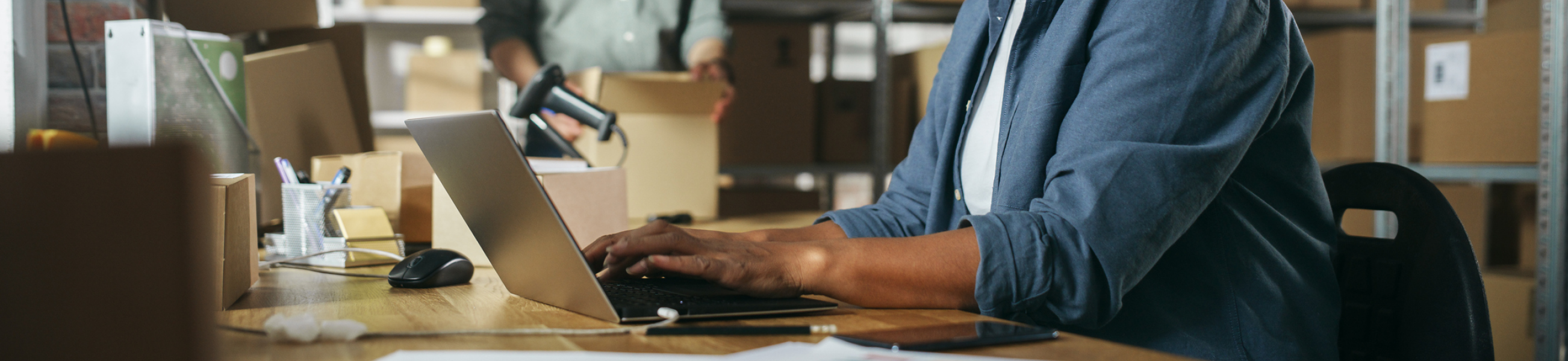 Woman in blue shirt typing on laptop on warehouse, with boxes on shelves behind him