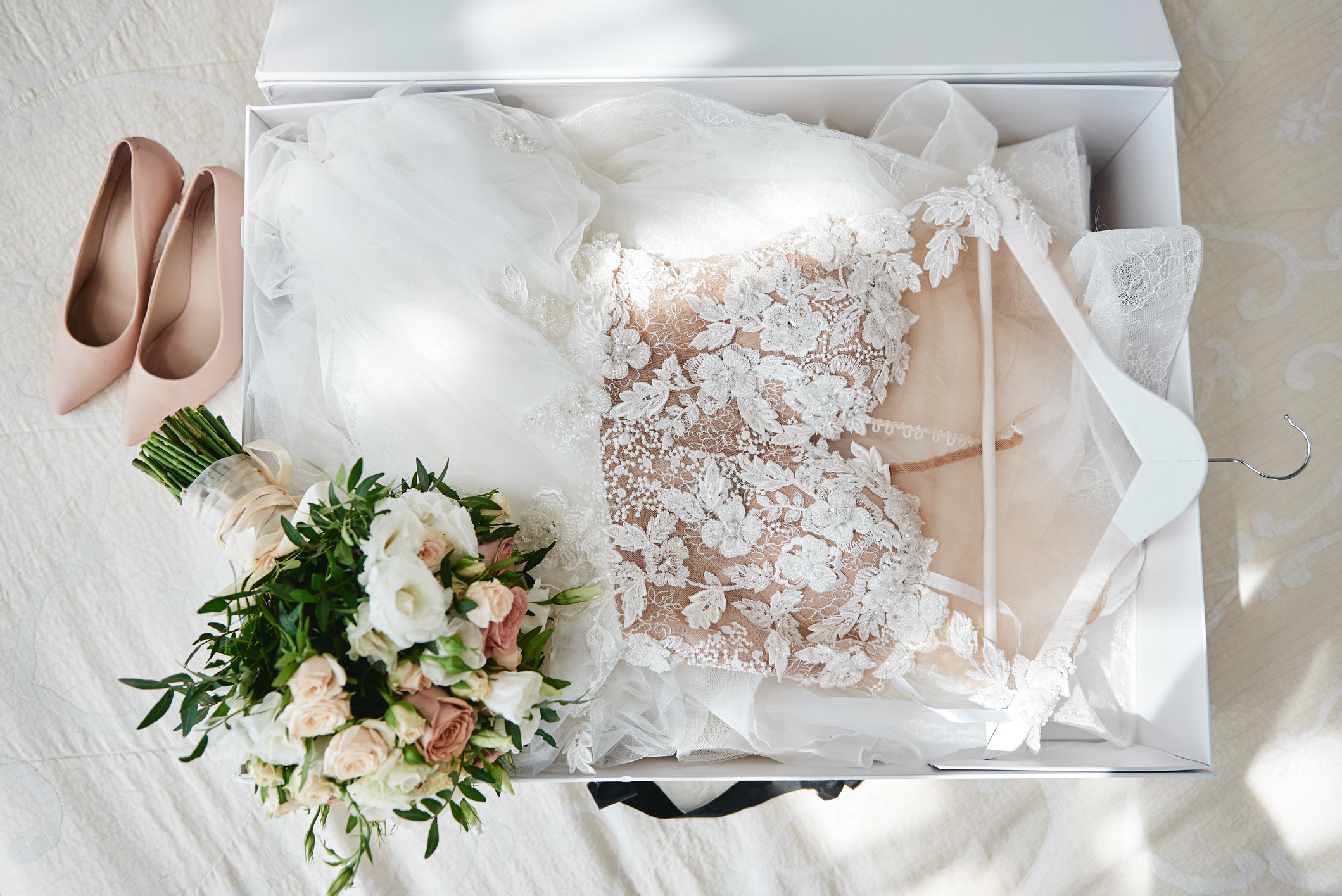Wedding dress in white delivery box, bridal bouquet resting on top and pale pink heels set to the side.