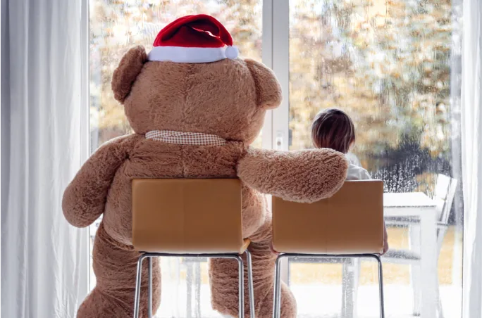 A little girl and big teddy bear in a Satna hat looking out the window