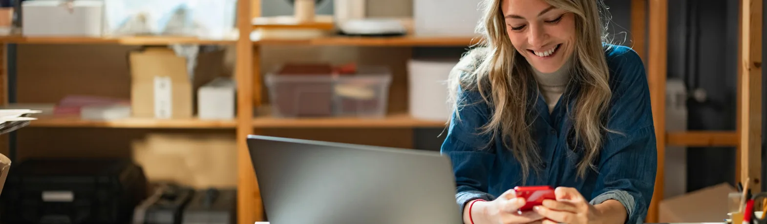 Female presenting person looks down at her phone and laptop in a room where she is operating her small business.