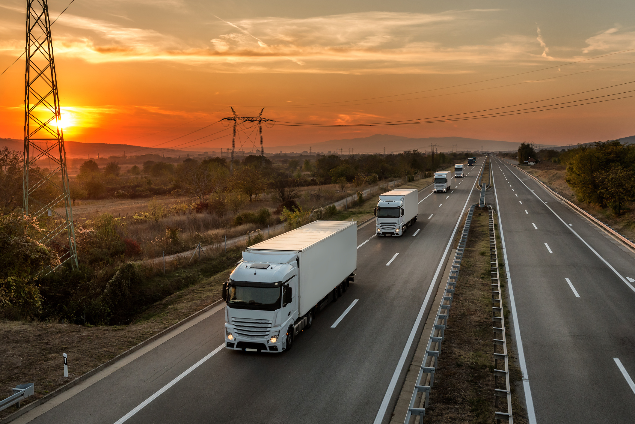 Unidentified white lorries driving down a country highway at sunrise.
