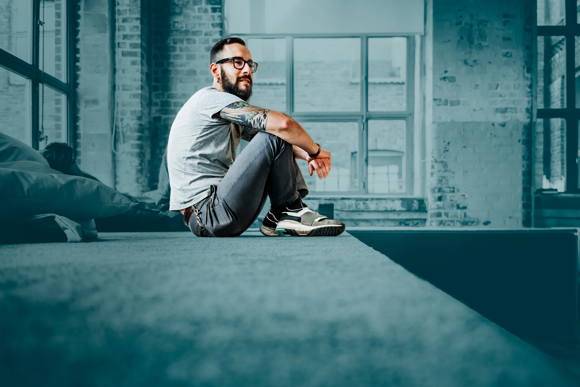 Een man die nonchalant op een verhoogd platform zit in een ruime kamer in industriële stijl, glimlachend naar de zijkant kijkt, met grote ramen en zichtbare bakstenen muren op de achtergrond.