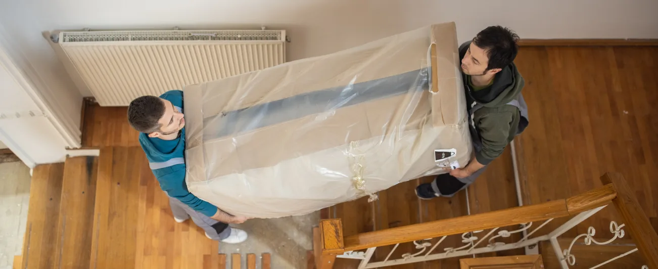 2 men carrying a large piece of packaged furniture up some wooden stairs