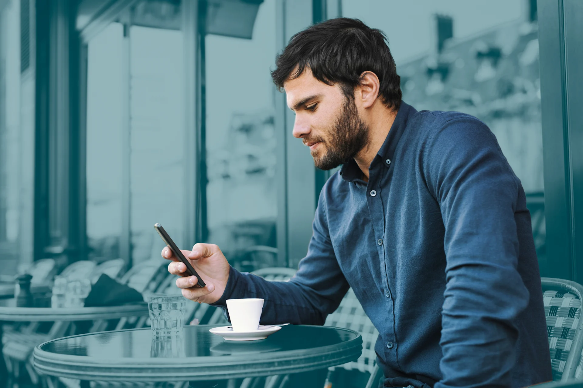 Een man met donker haar en een baard, gekleed in een donkerblauw overhemd, zit aan een tafeltje op een terras. Hij kijkt naar beneden en houdt een smartphone in zijn handen. Een klein wit espressokopje met schoteltje staat op de tafel voor hem. De afbeelding heeft een turquoise kleuroverlay.