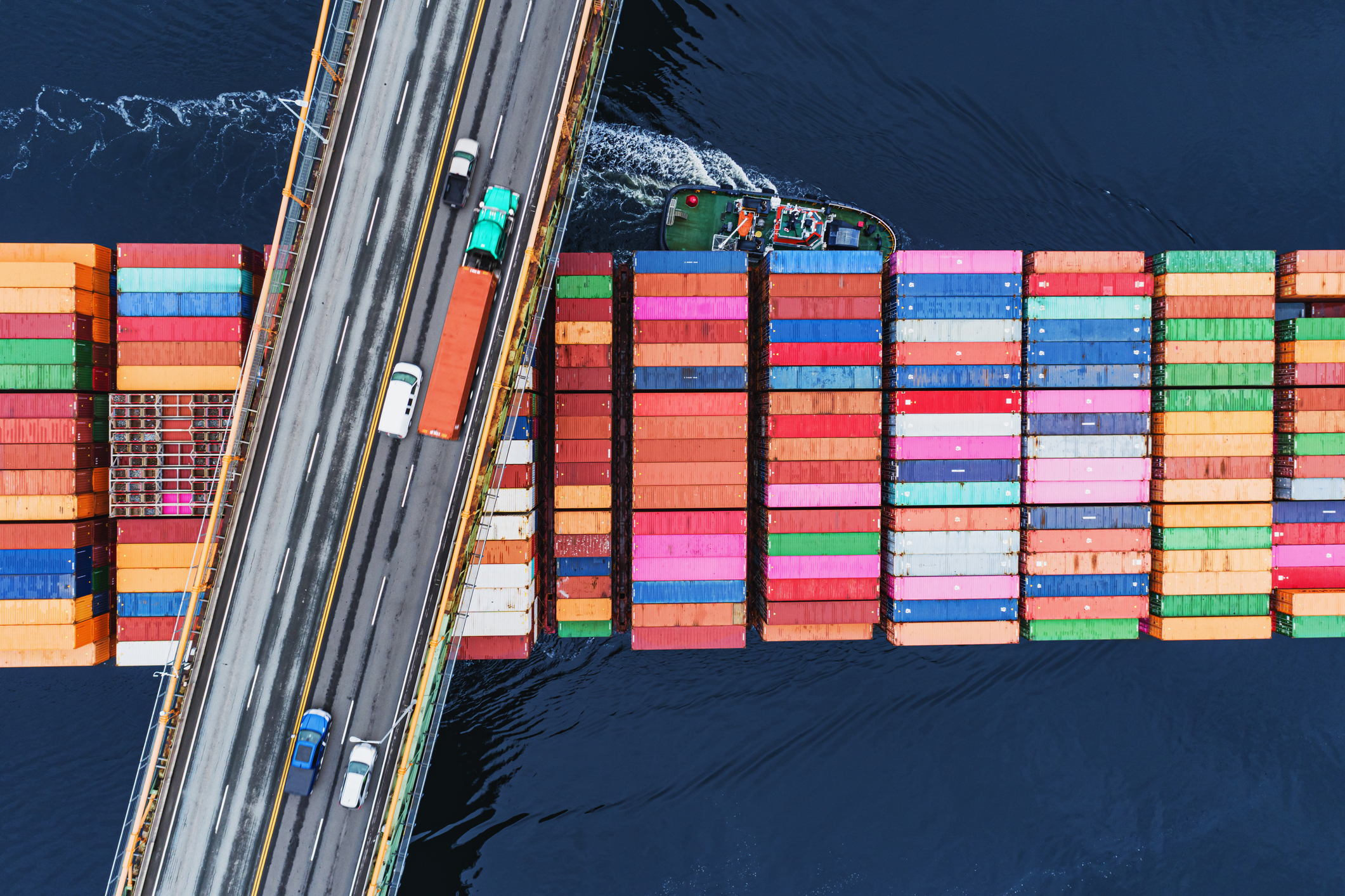 A cargo boat stacked with colourful shipping containers passes beneath a bridge road.