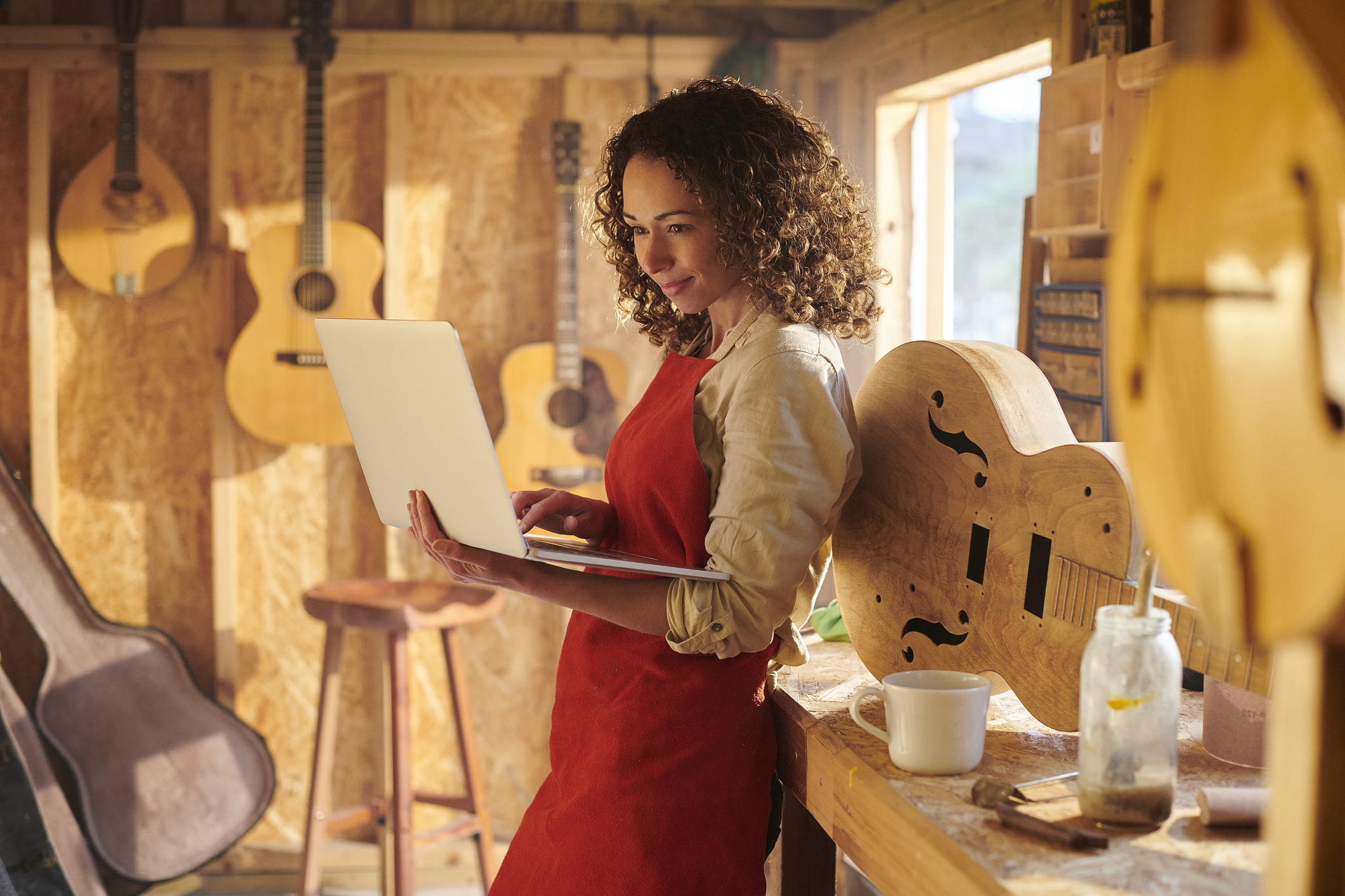 Female presenting person in a woodshop, making instruments, leans on a table and works on a laptop.