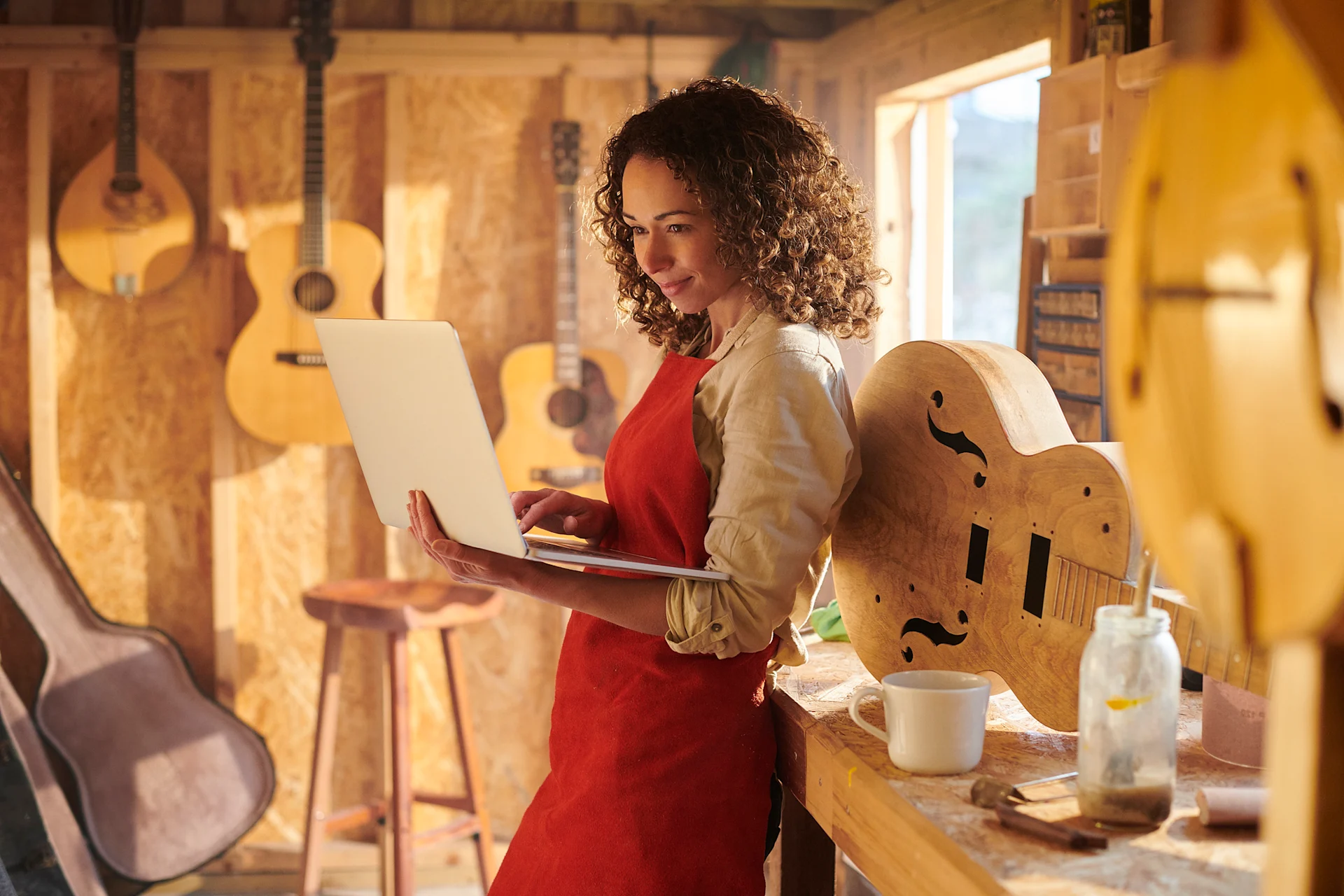 Female presenting person in a woodshop, making instruments, leans on a table and works on a laptop.