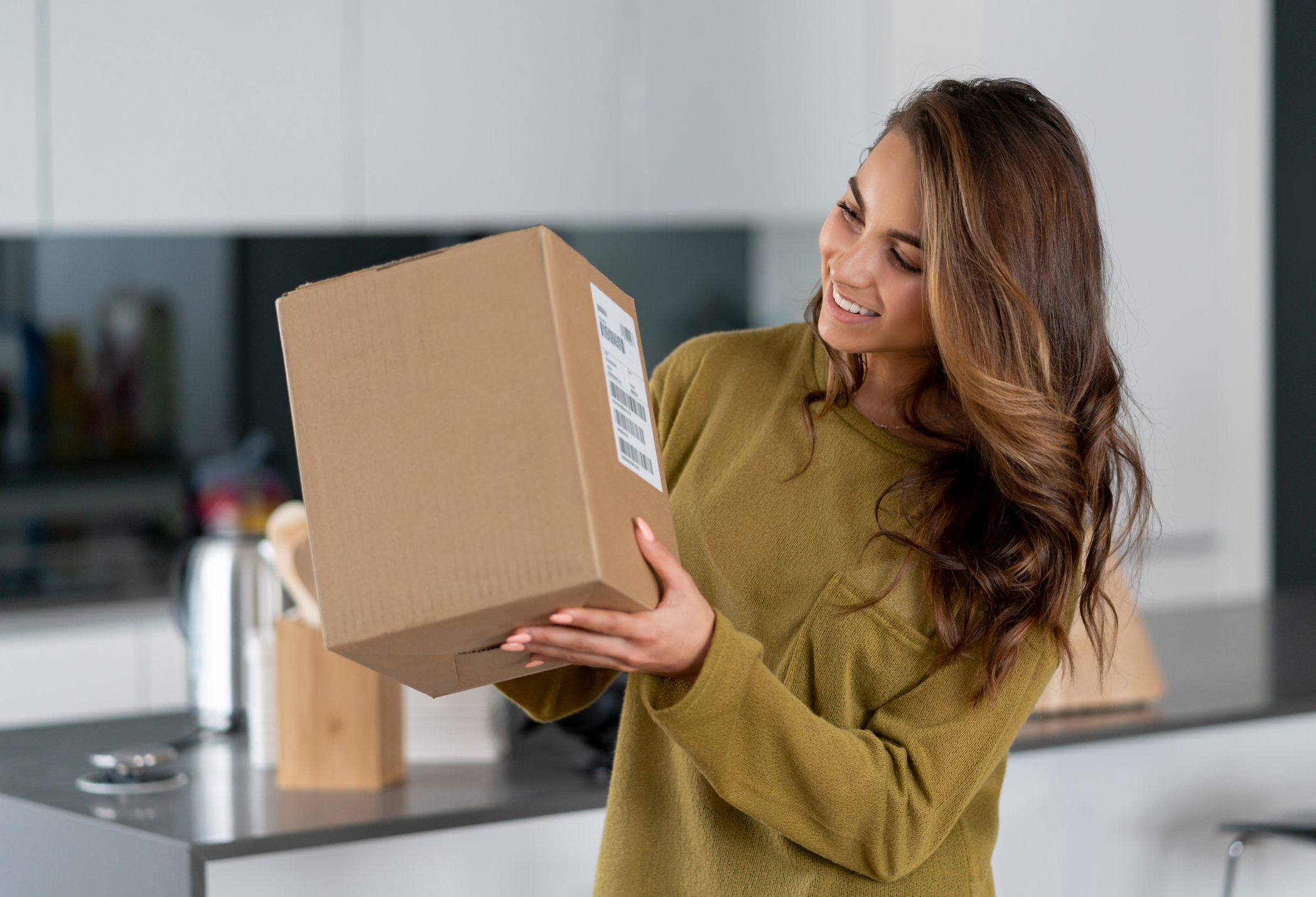 Female presenting person smiling at parcel box in their kitchen.