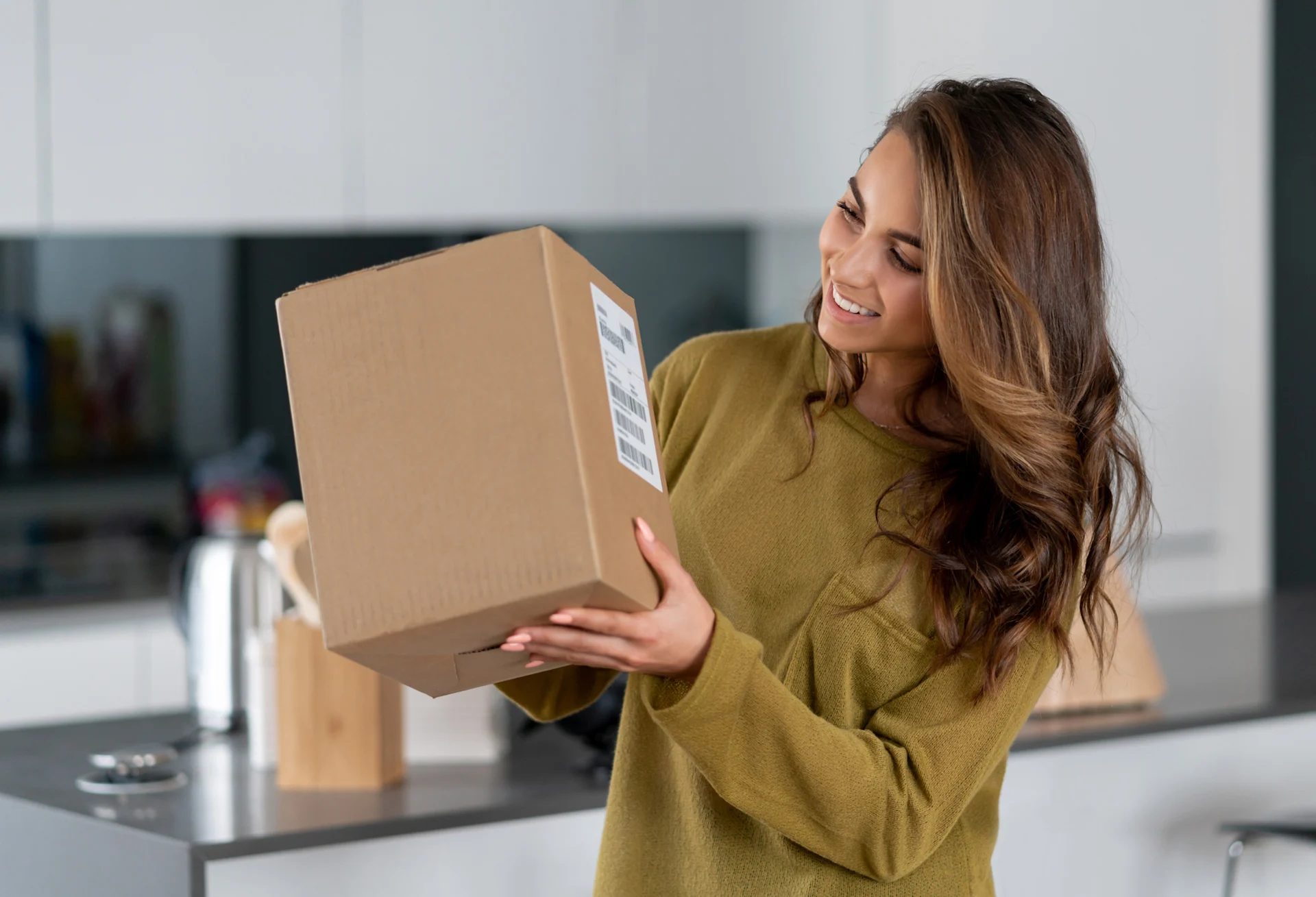 Female presenting person smiling at parcel box in their kitchen.