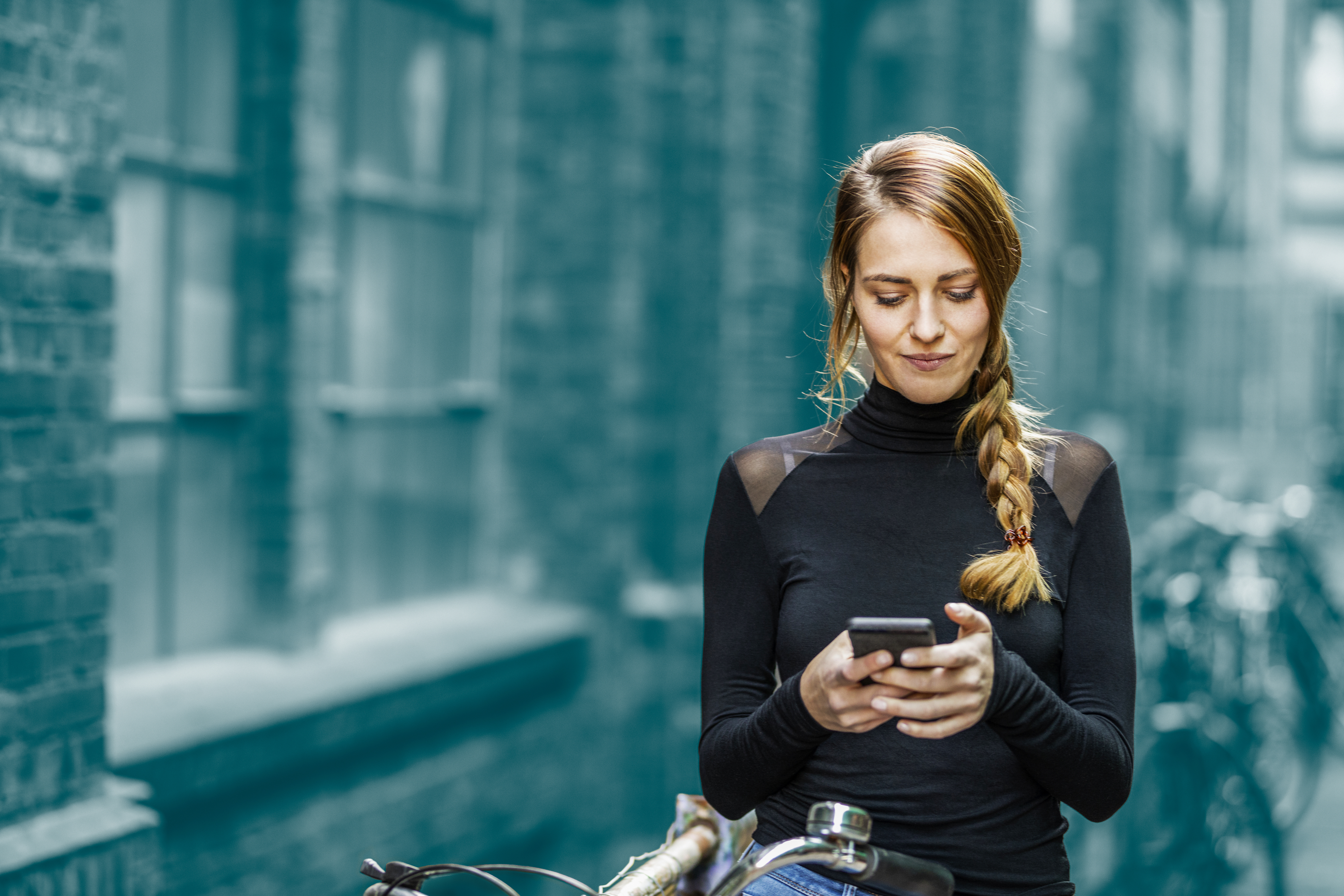 A woman with a long side braid stands beside a bicycle in an urban alley, looking down at her smartphone. She is wearing a black long-sleeve top, and brick buildings with windows blur into the background.